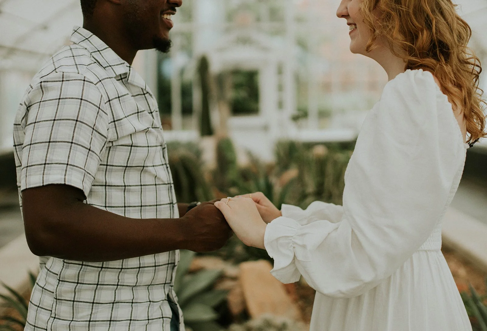 A couple holding hands and smiling at each other indoors with plants and a window in the background.