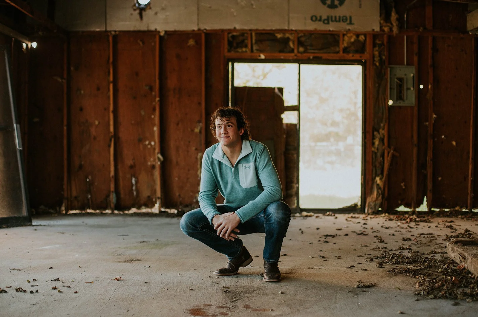 Young man with curly hair squatting in an abandoned, unfinished room with dirt and debris on the floor and partially exposed wooden walls.