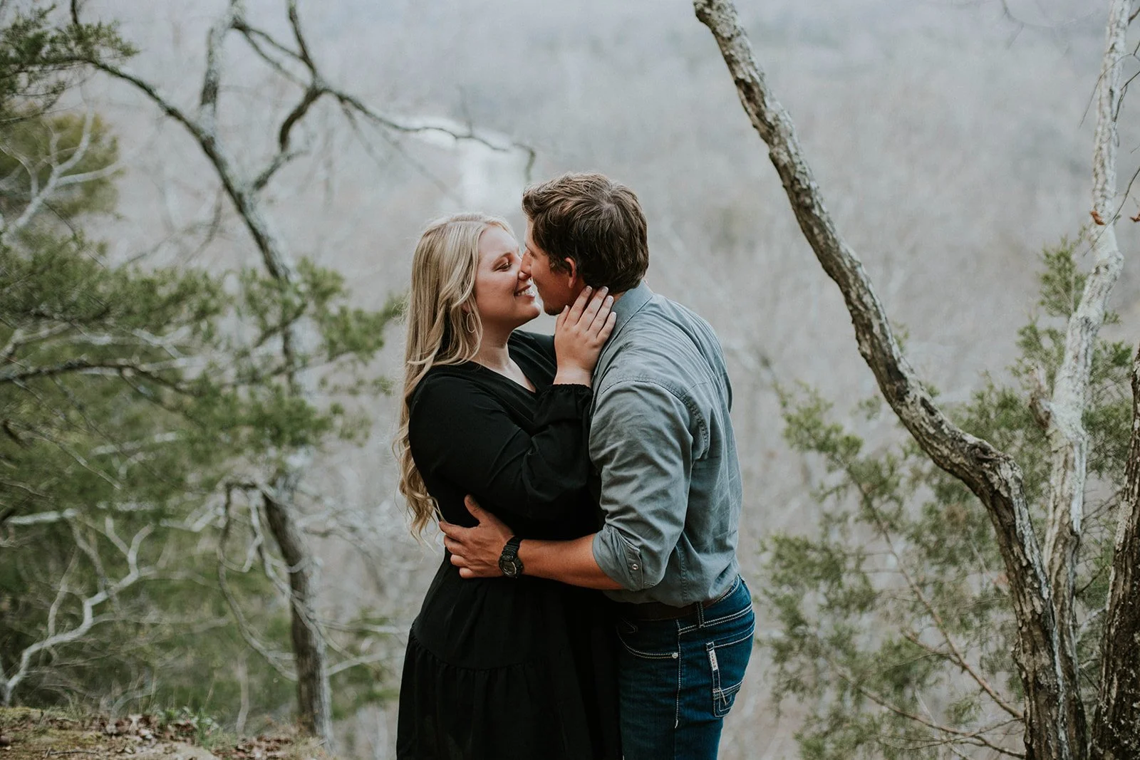 A couple is about to kiss outdoors among trees and foliage, with the woman wearing a black dress and the man in a blue shirt and jeans.