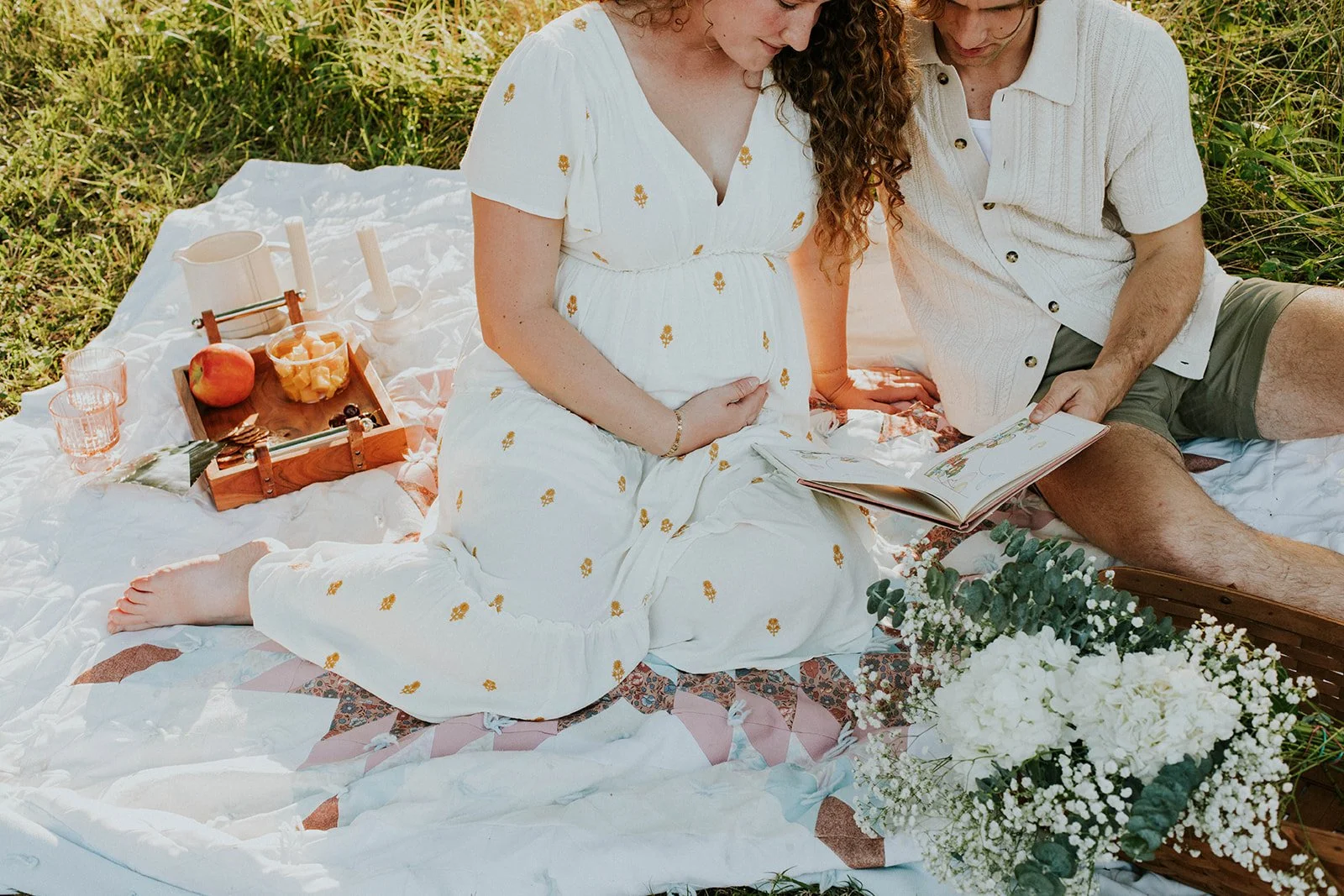 A pregnant woman and a man sitting on a blanket during a picnic in a grassy outdoor setting, reading a book together. The woman is wearing a white dress with small yellow flowers, and the man is wearing a cream shirt and shorts. There is a tray of sn