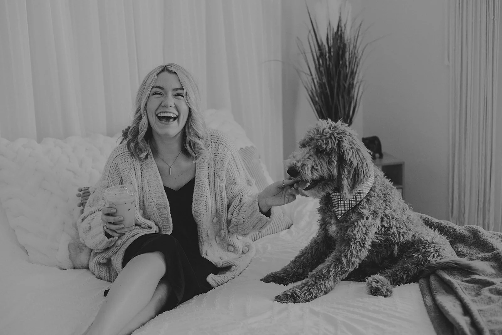 A woman laughing while petting a large curly-haired dog on a bed in a cozy room.