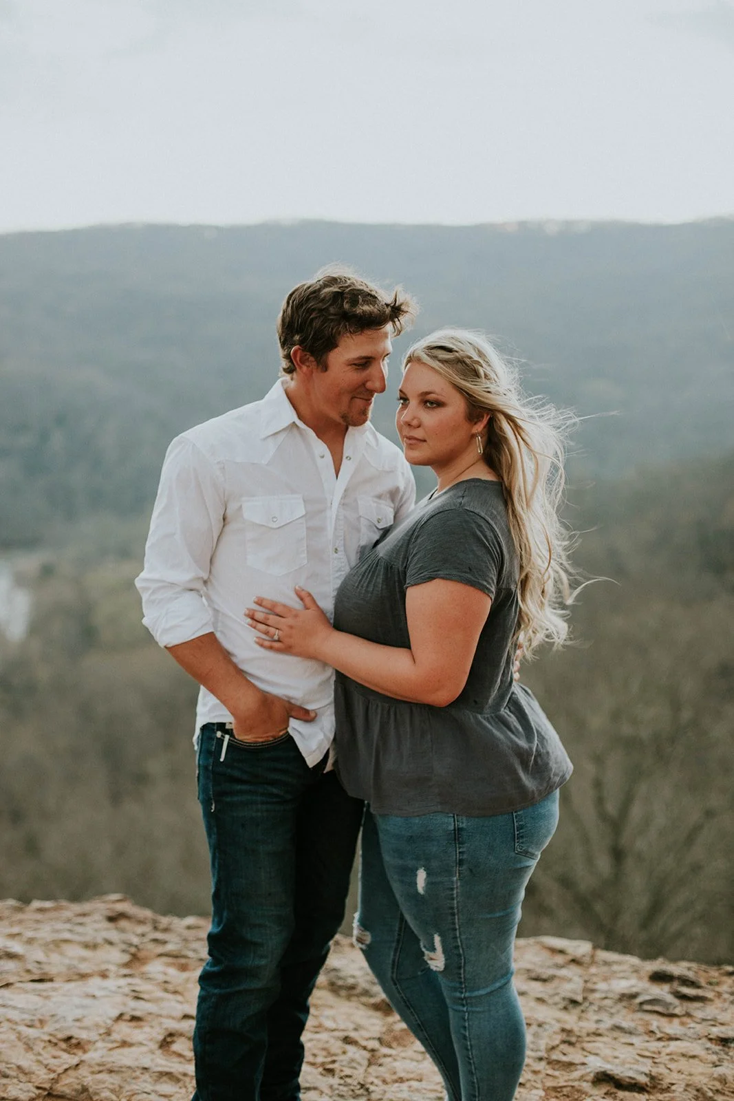 A couple standing close together outdoors with a scenic mountainous background, the man in a white shirt and the woman in a gray top.