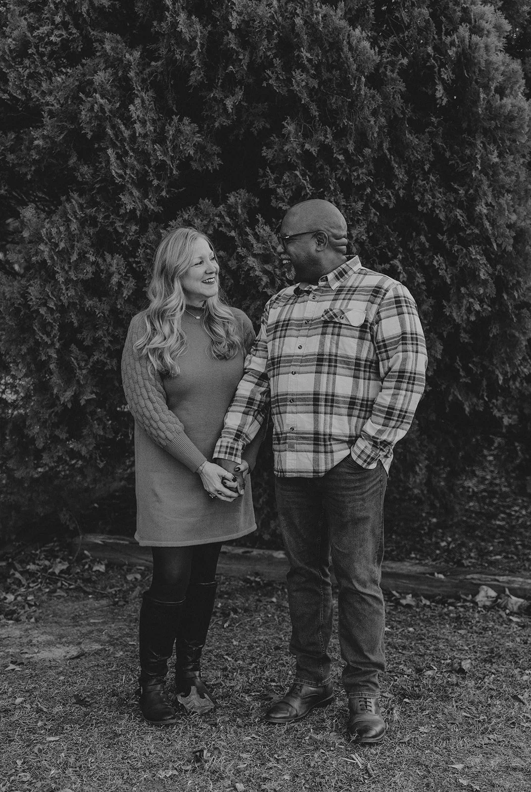A black and white photograph of a woman and man standing outdoors in front of a large tree, holding hands and smiling at each other.