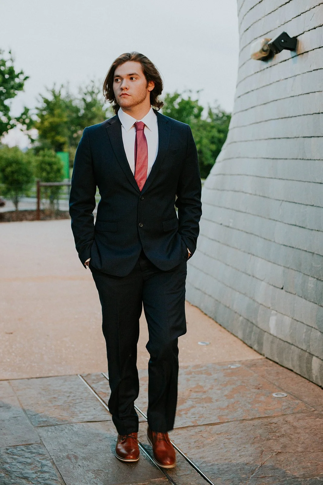 A young man in a black suit, white shirt, red tie, and brown shoes walking outdoors near a curved white brick wall.