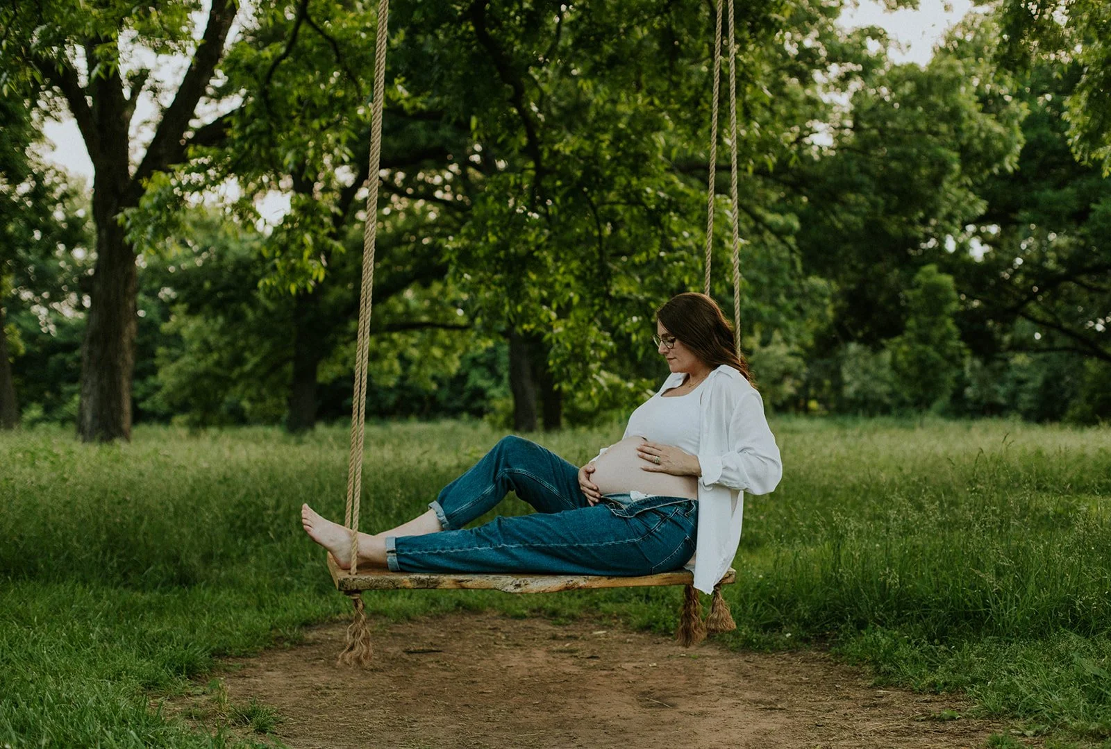 Pregnant woman sitting on a swing in a park, gently touching her belly with both hands, surrounded by green trees.