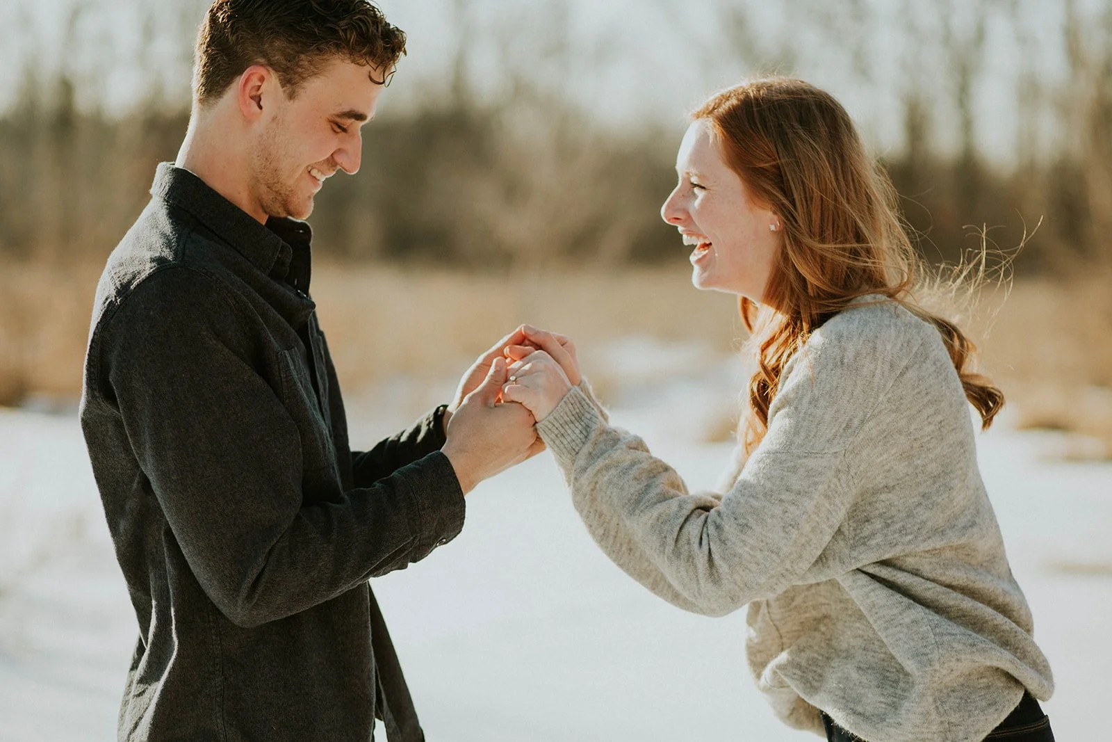 A young man and woman outside in the snow, holding hands and smiling at each other, enjoying a happy moment.