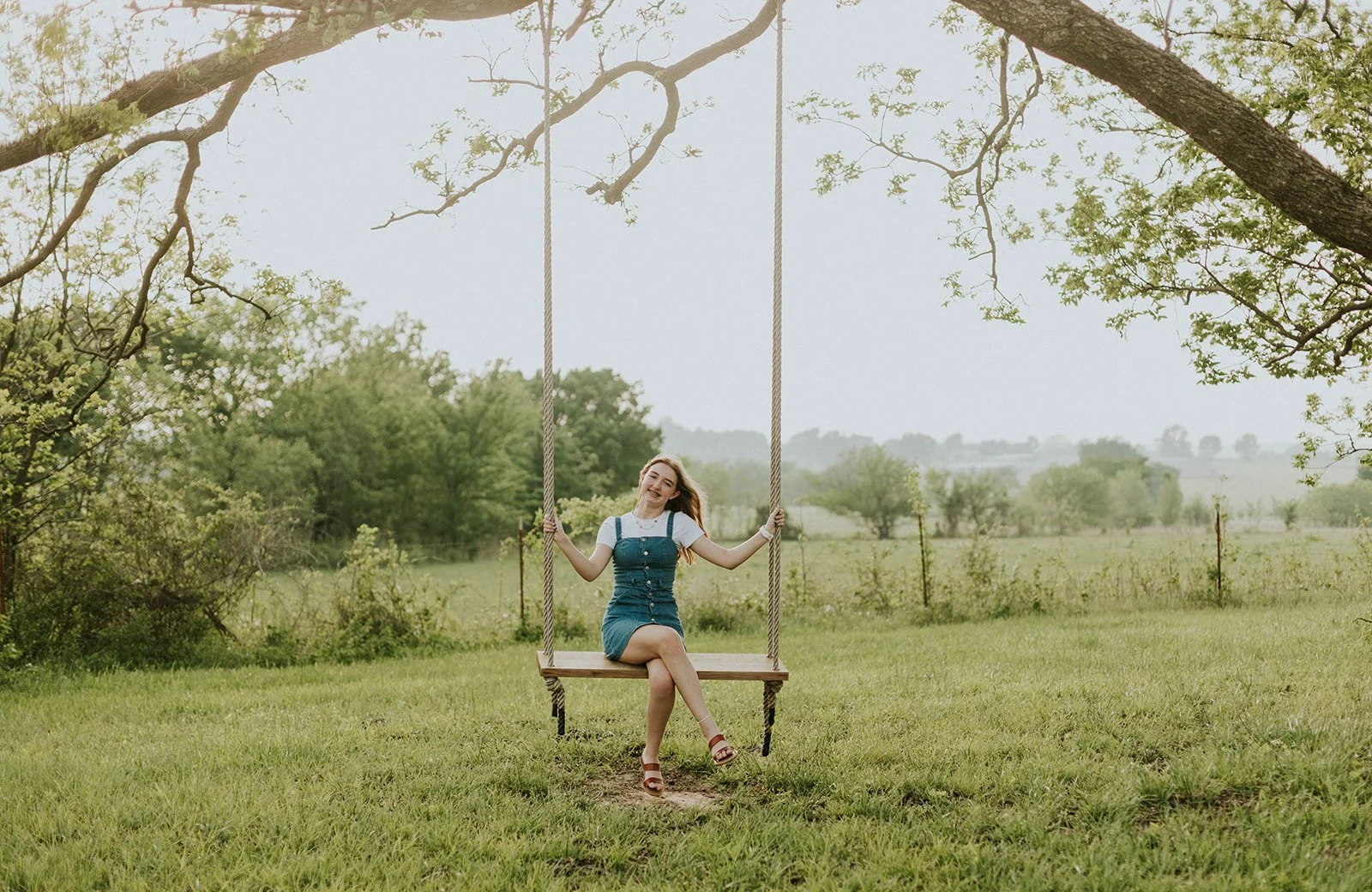 A young woman with long hair sitting on a wooden swing hanging from a tree in a grassy field, smiling, with a scenic background of trees and open land.