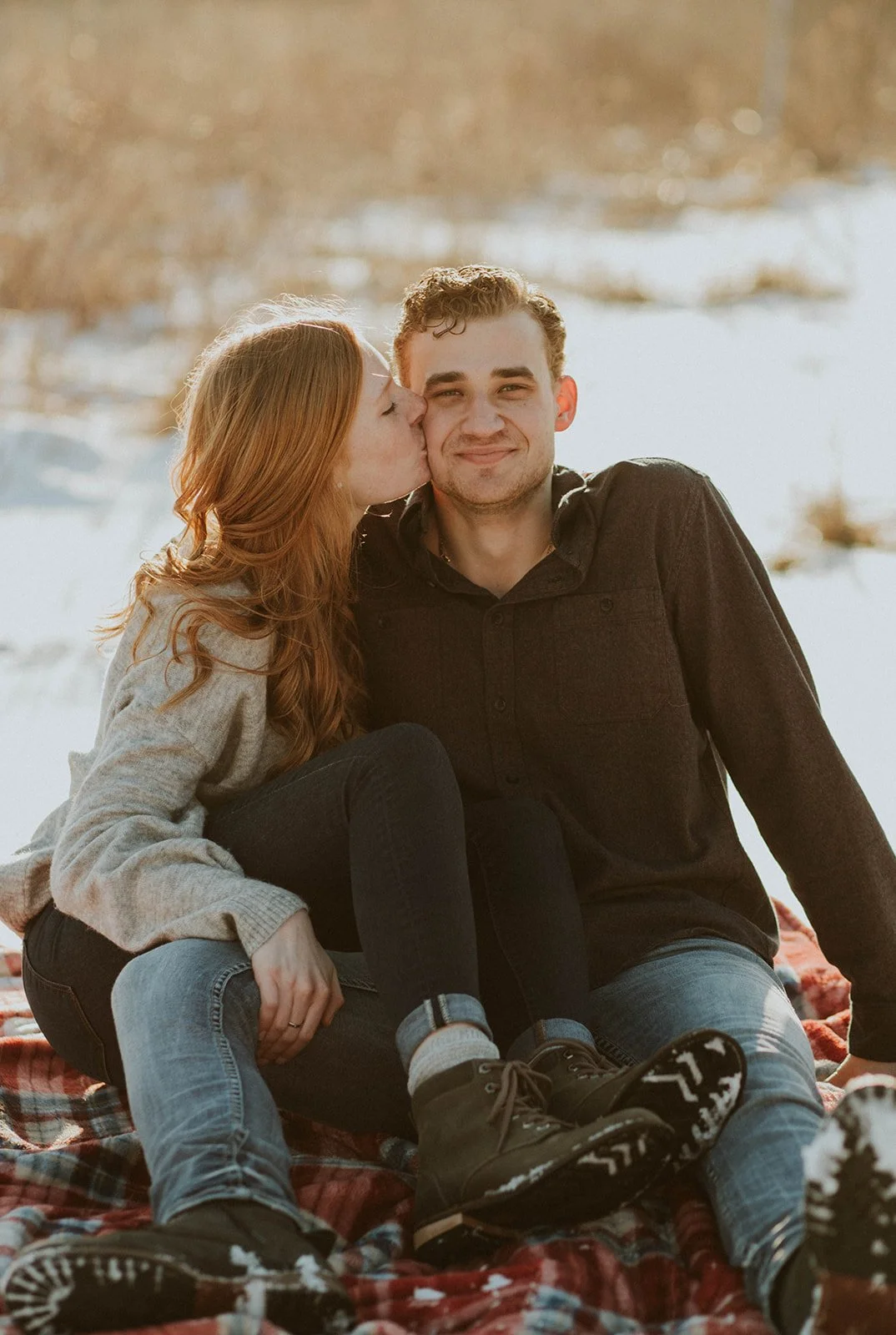 A couple sitting on a blanket outdoors, with the woman giving the man a kiss on the cheek, both smiling, near a body of water.