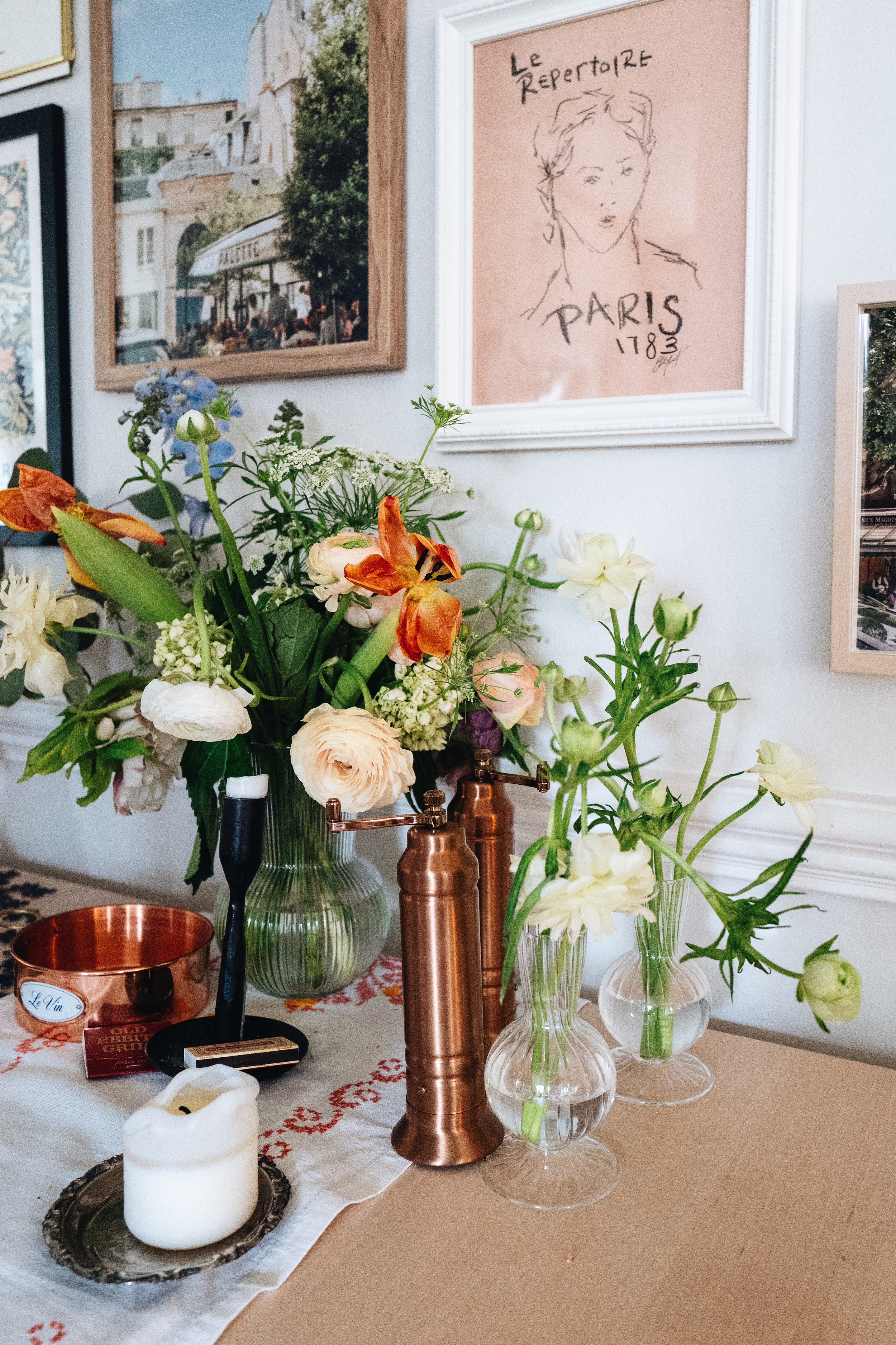 Pink, blue, and white flowers on a dining room table with copper salt and pepper shakers and a pillar candle. In front of a gallery wall.