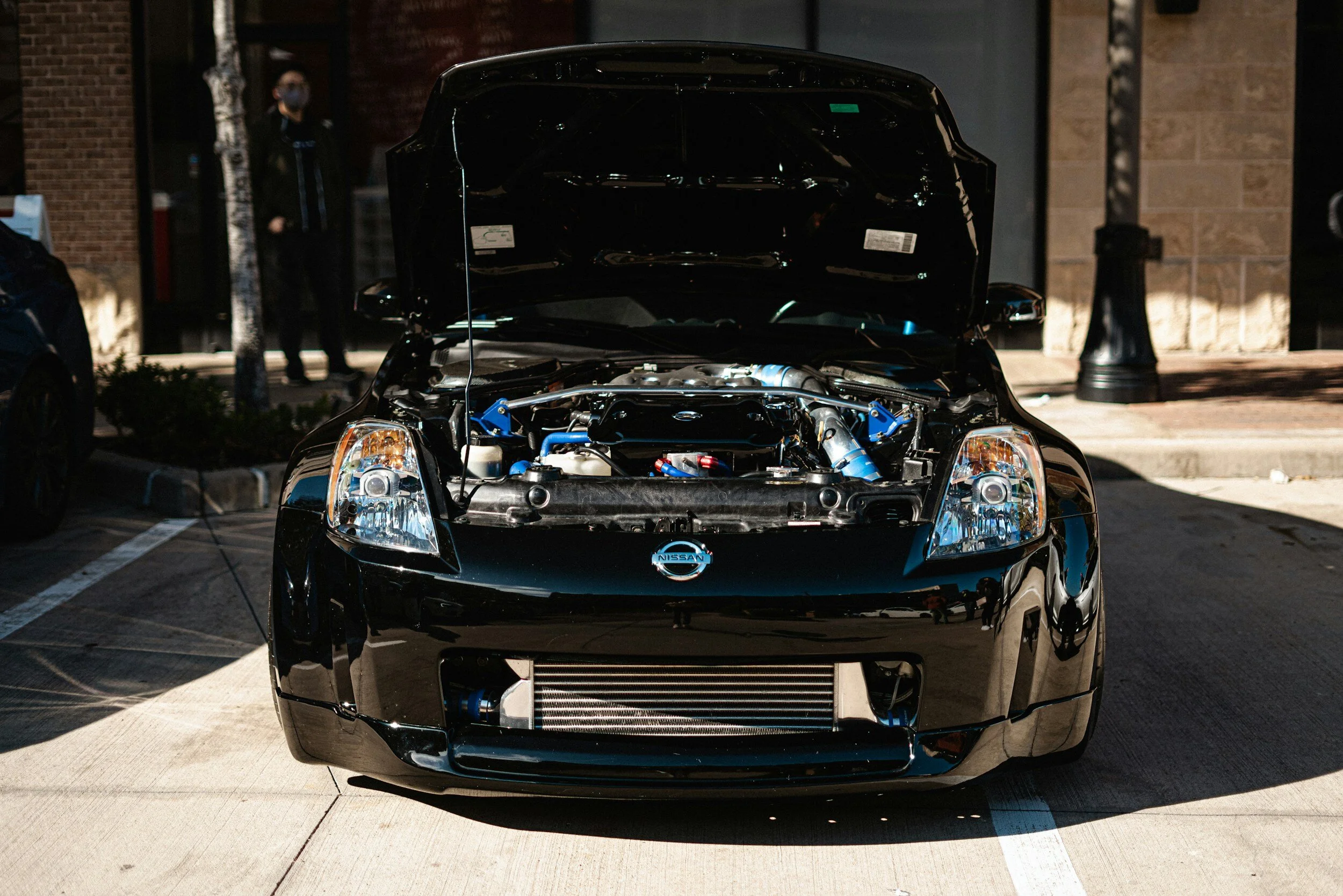A black Nissan sports car with the hood open, showing the engine. Parked on a street, with a blurred building and person in the background.