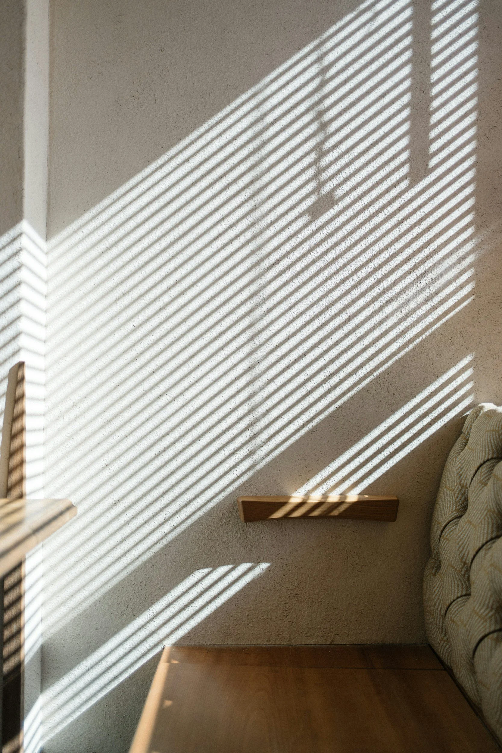 Sunlight casting diagonal striped shadows through window blinds onto a beige wall in a room with a wooden surface and a patterned couch.
