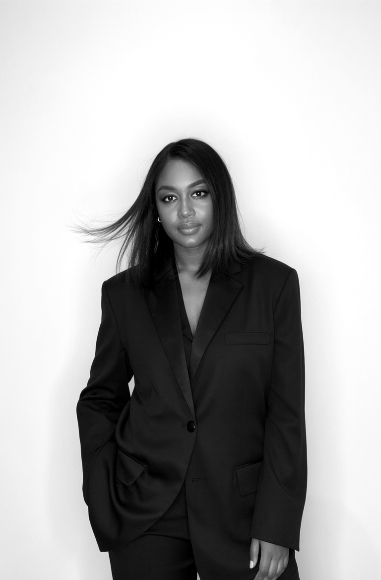 A young woman with medium-length black hair and earrings, wearing a black blazer and pants, stands against a plain white wall, looking directly at the camera.