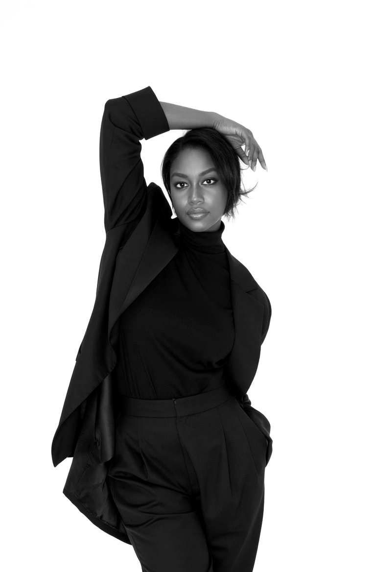Black and white portrait of a woman in a black blazer, turtleneck, and pants, standing against a plain white background with one hand resting on her head.