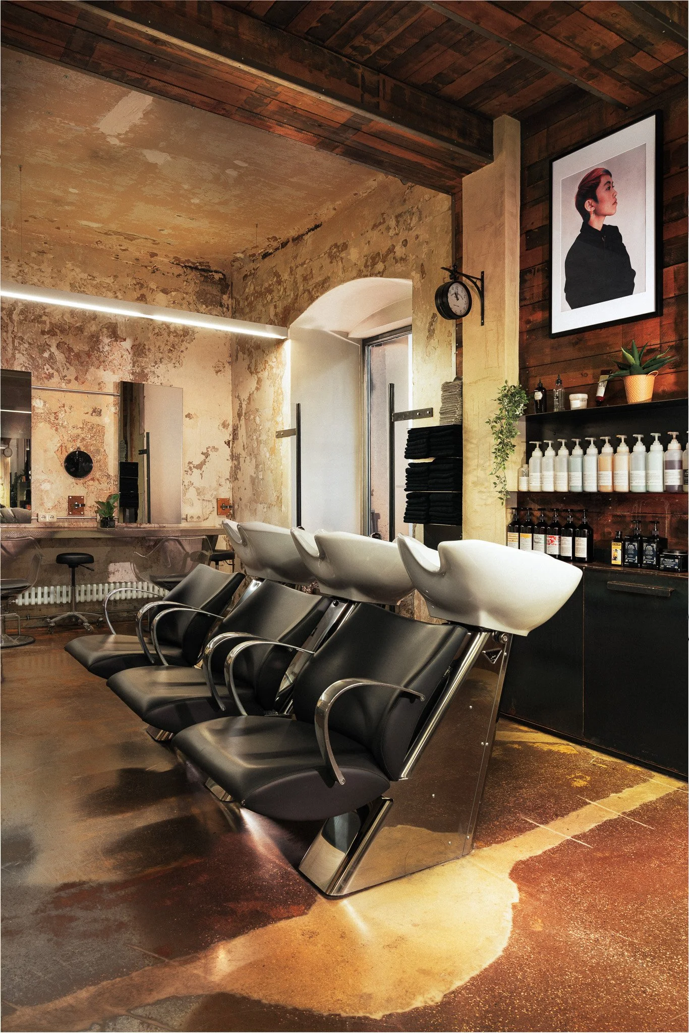 Ein Friseursalon mit drei schwarzen und drei weißen Waschkabinen, schwarzen Stühlen, an der Wand sind Handtücher, Produkte und ein Bild einer Frau, die Haare hat. Das Interieur kombiniert eine alte, beschädigte Wand mit moderner Inneneinrichtung.