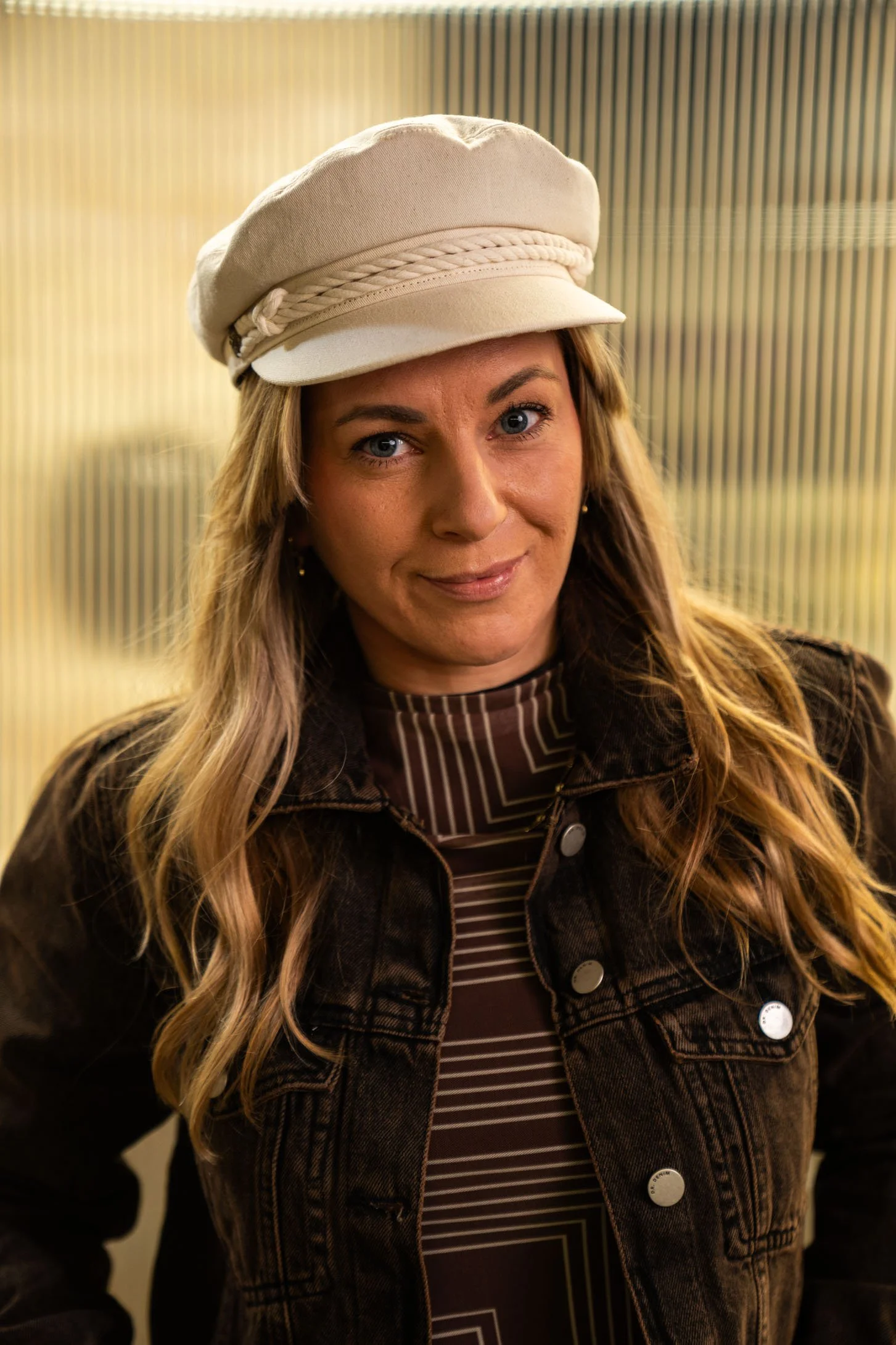 Eine Frau mit blonden, gewellten Haaren trägt einen weißen Hut und eine schwarze Jeansjacke, steht vor einem Hintergrund mit senkrechten Linien in Grau und Beige.