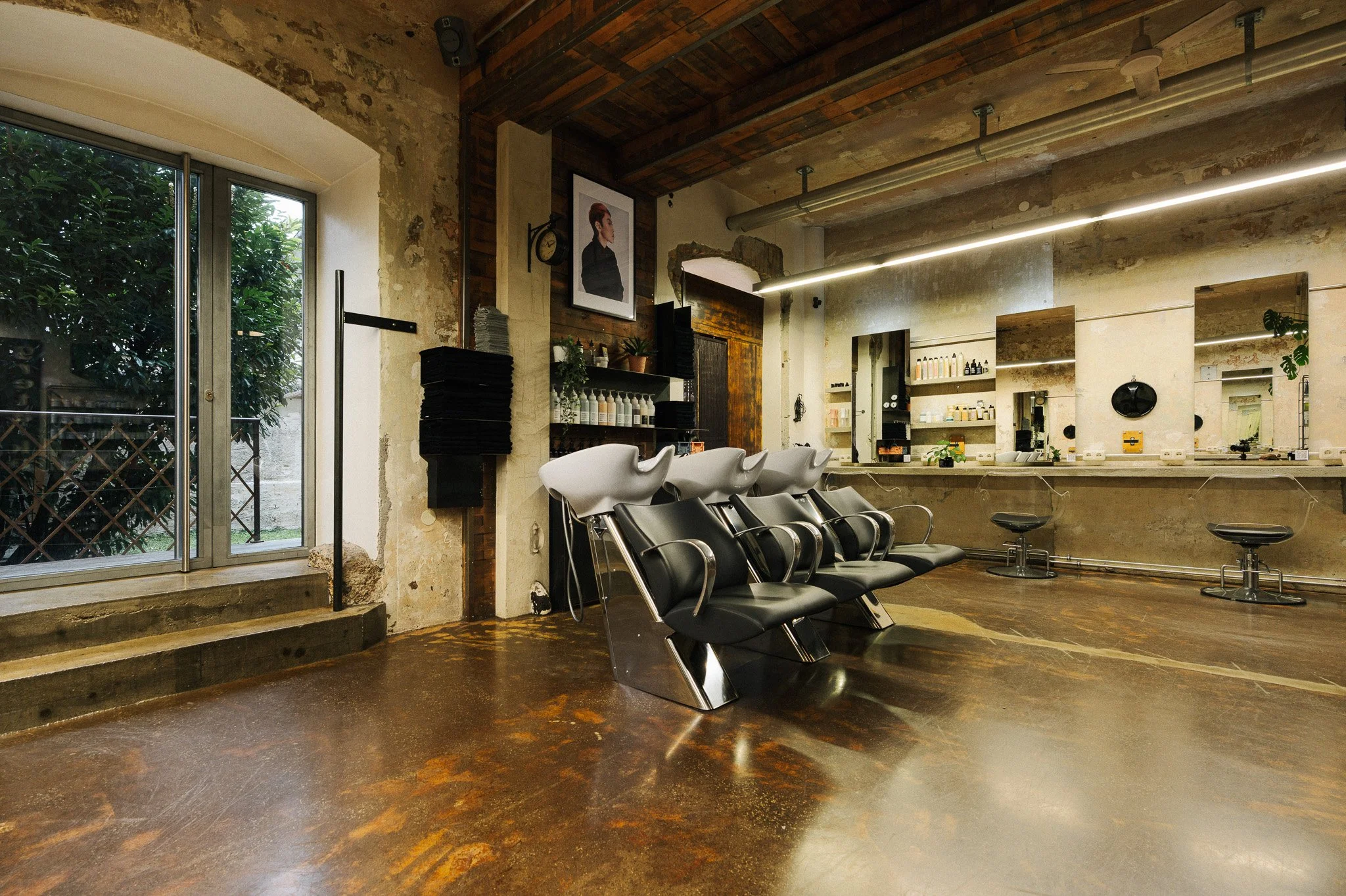 Innenansicht eines modernen Friseursalons mit Waschbecken, schwarzen Stühlen, Spiegeln und Produkten auf Regalen, große Fenster mit Blick nach draußen.