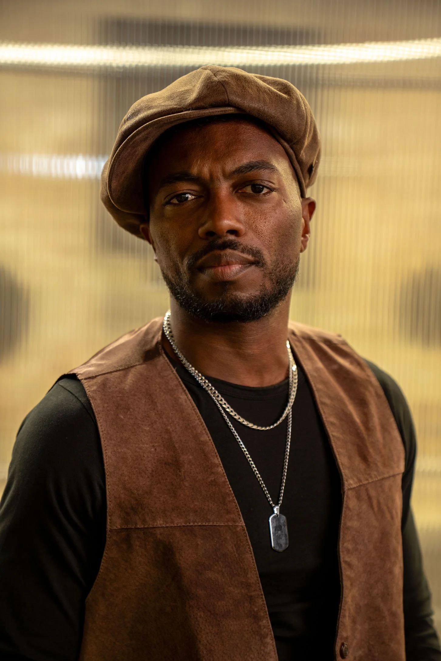 Porträt eines Mannes mit dunklem Teint, schwarzes T-Shirt, brauner Weste, Lederkette und Dog Tag, im Hintergrund ausgeleuchtete, strukturierte Wand.