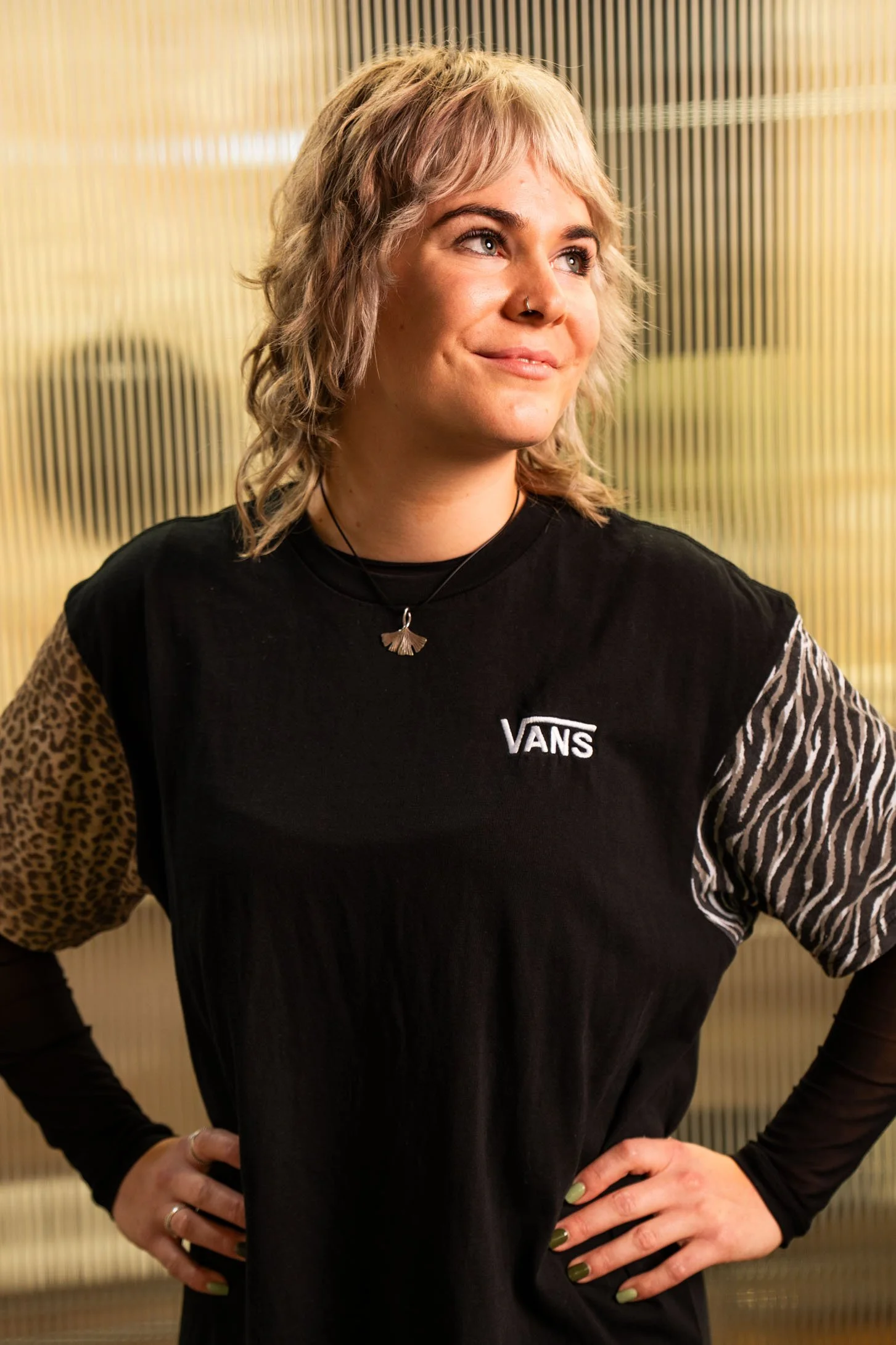 Junge Frau mit blondem, lockigem Haar, trägt ein schwarzes T-Shirt mit Leoparden- und Zebrastreifenärmeln, steht vor einem gelben Muster-Hintergrund.