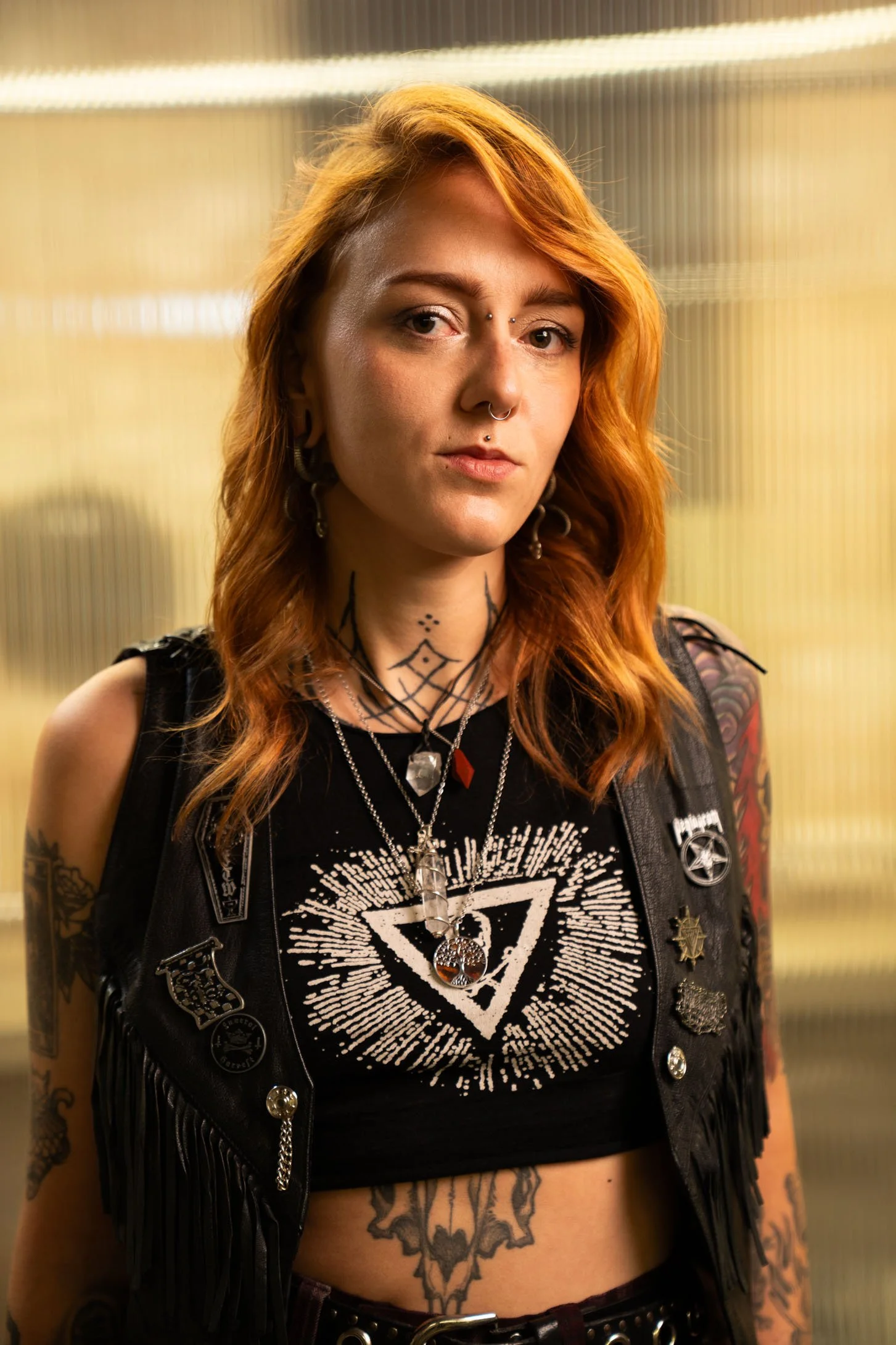 Junge Frau mit roten, Lockigen Haaren und Tattoos, trägt schwarze Band-T-Shirt mit weißem Design, schwarze Lederjacke mit Abzeichen und Ketten, im Hintergrund eine glasartige Wand