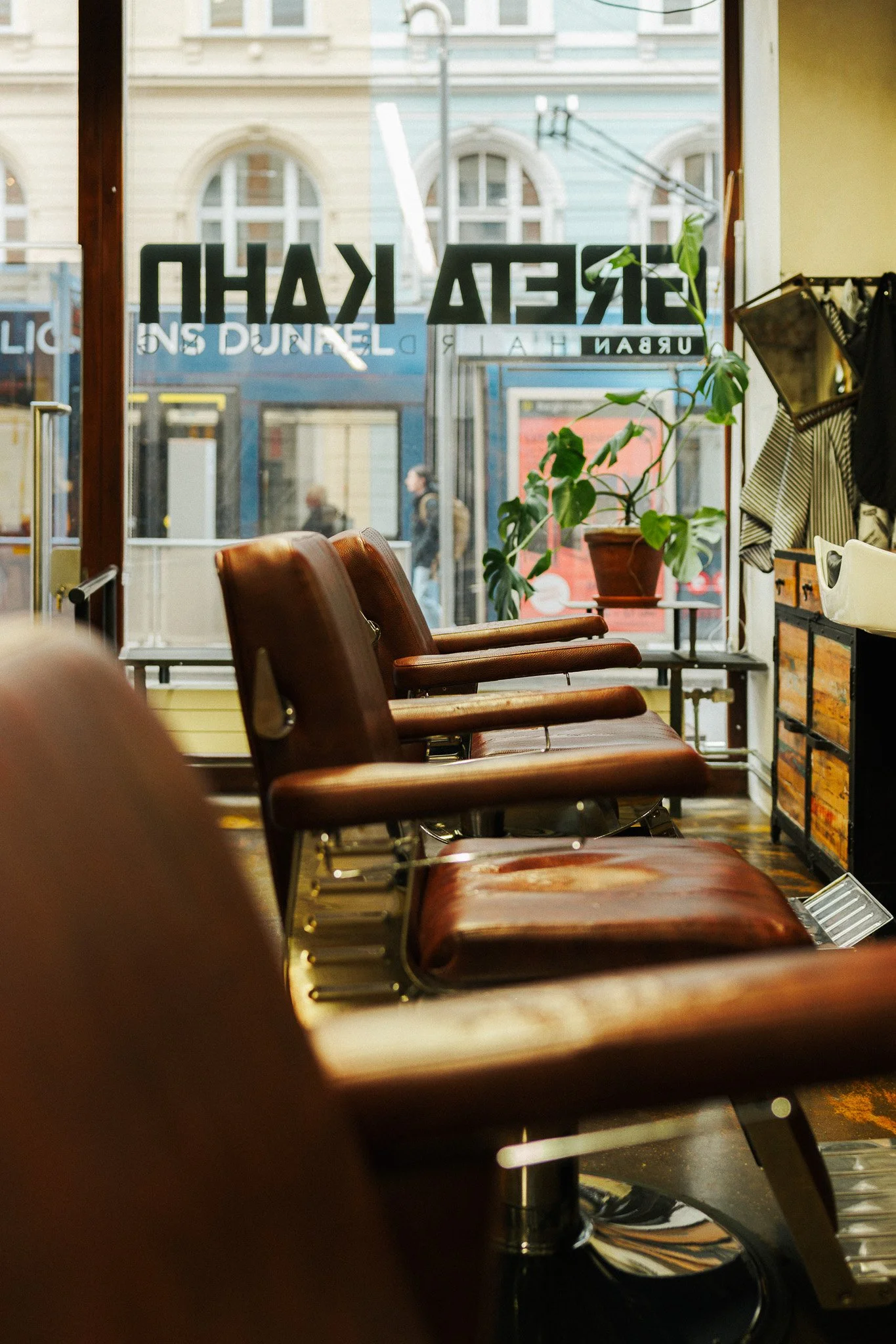 Leere braune Friseurstühle vor einem großen Fenster mit Blick auf die Straße, innen mit Pflanzen und Dekorationen.