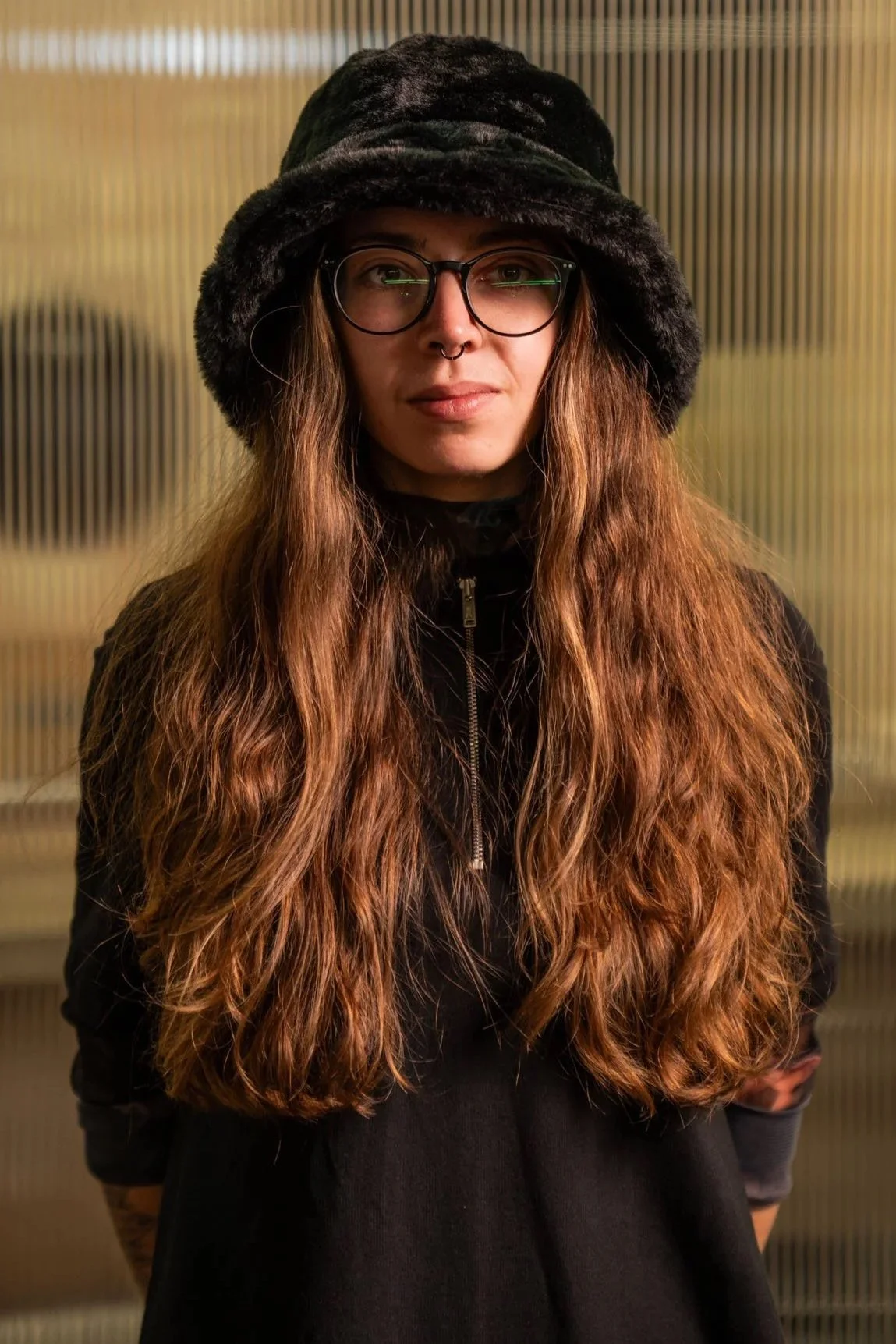 Jugendliche Frau mit langen, lockigen braunen Haaren, schwarzen Brillengläsern, Nasenpiercing und schwarzem Hut, trägt schwarze Jacke, steht vor einer strukturierten Hintergrundwand.