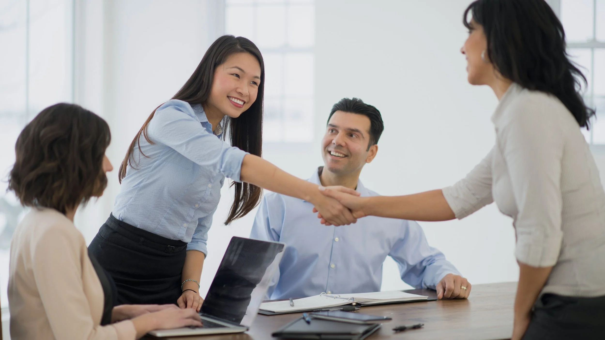 A group of five people in a business meeting, with two women shaking hands, one woman seated with a laptop, and others observing and smiling in a bright office setting.