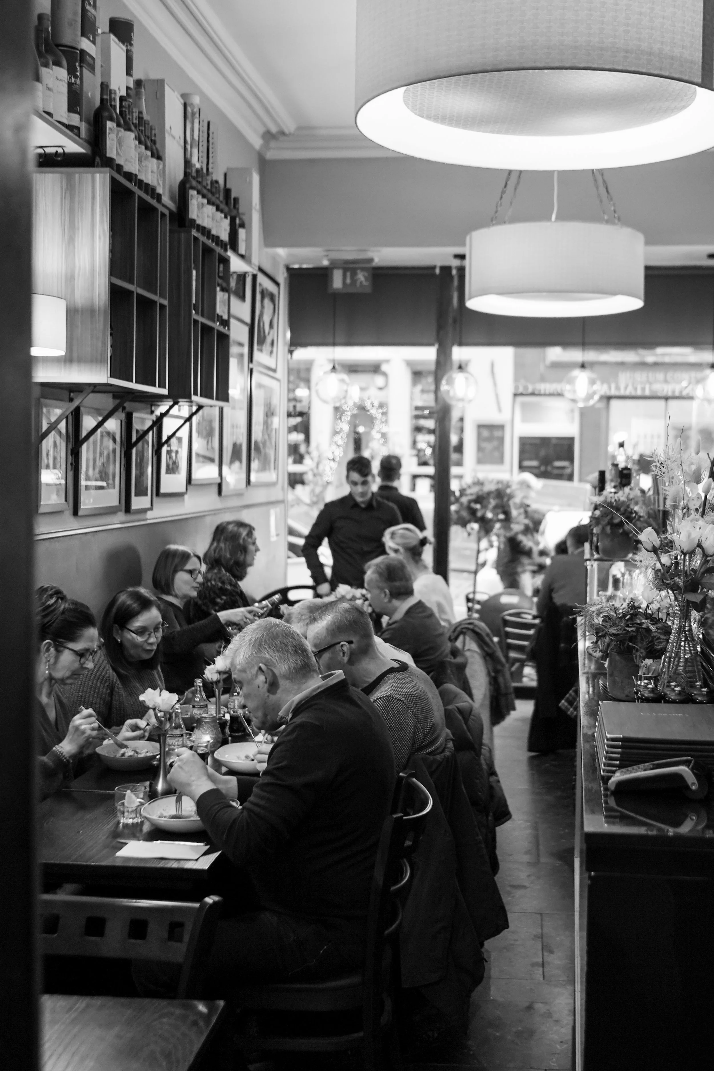 Black and white photo of a restaurant interior with people dining at a long table, some of them eating and talking, with waiter serving in the background, and framed pictures on the wall.