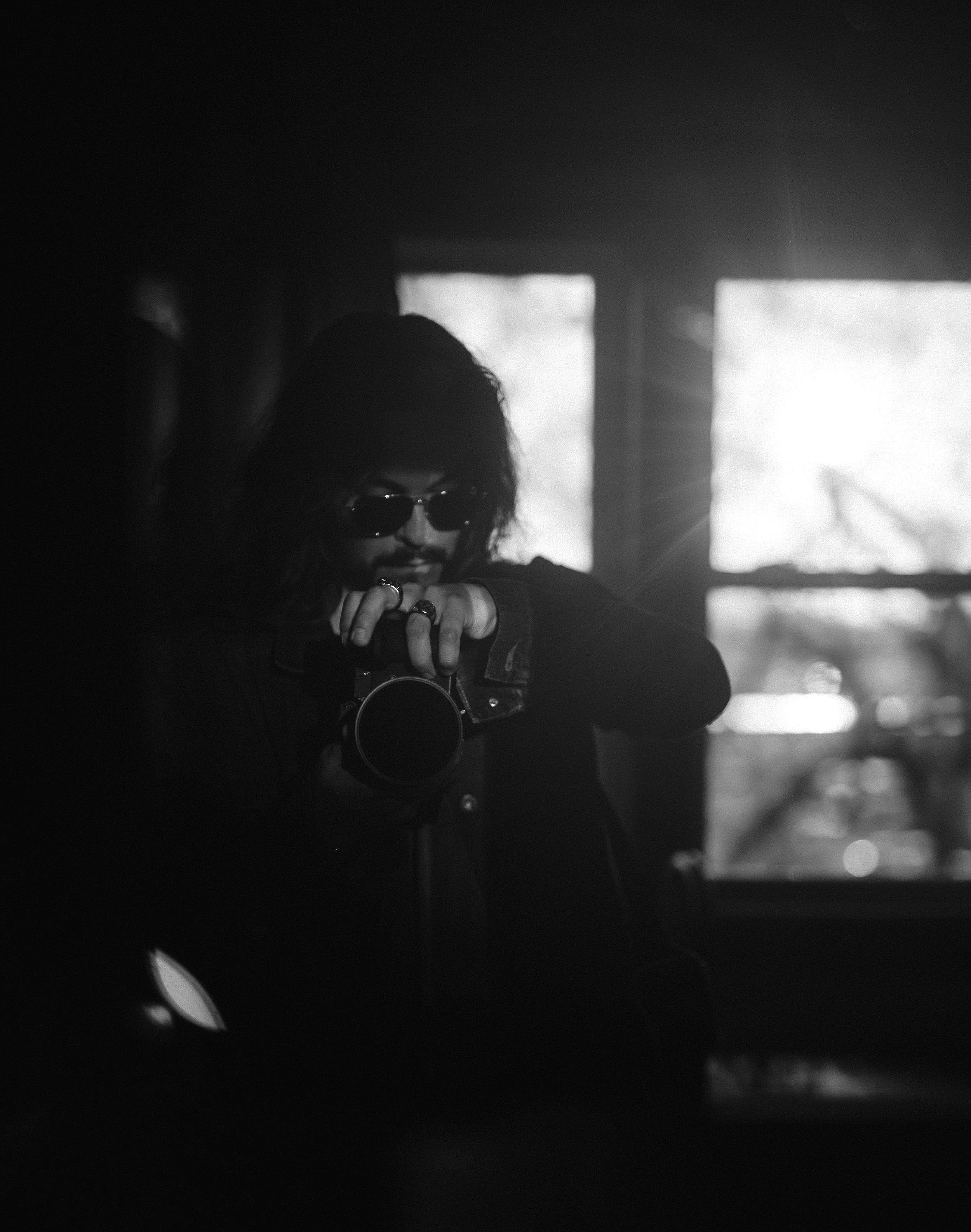 A person with sunglasses is looking down and smiling, with sunlight coming through a window in the background.