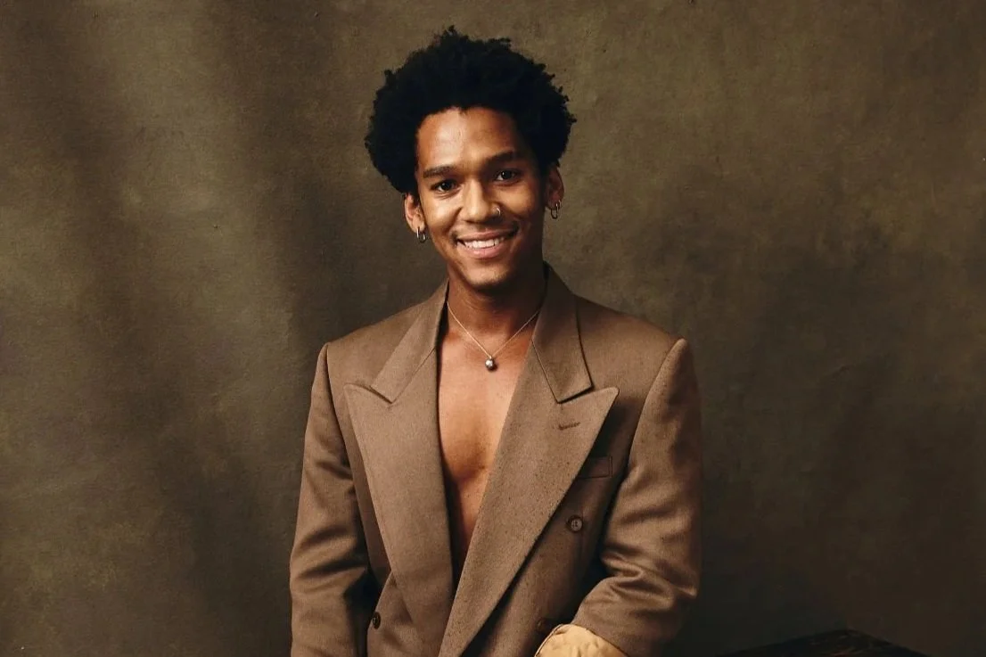 Young man with curly hair, wearing a brown blazer and jewelry, smiling at the camera in front of a plain brown background.