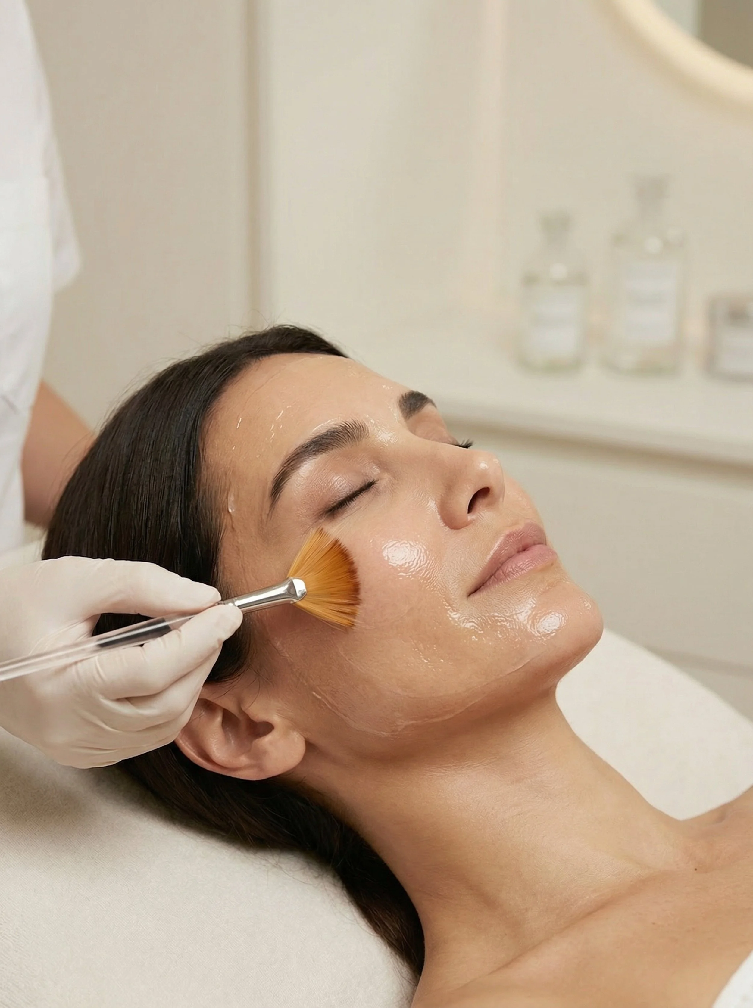A woman receiving a facial  chemical peel treatment, with a clinician applying a facial product with a brush, while holding a small glass bowl and a bottle of facial serum.