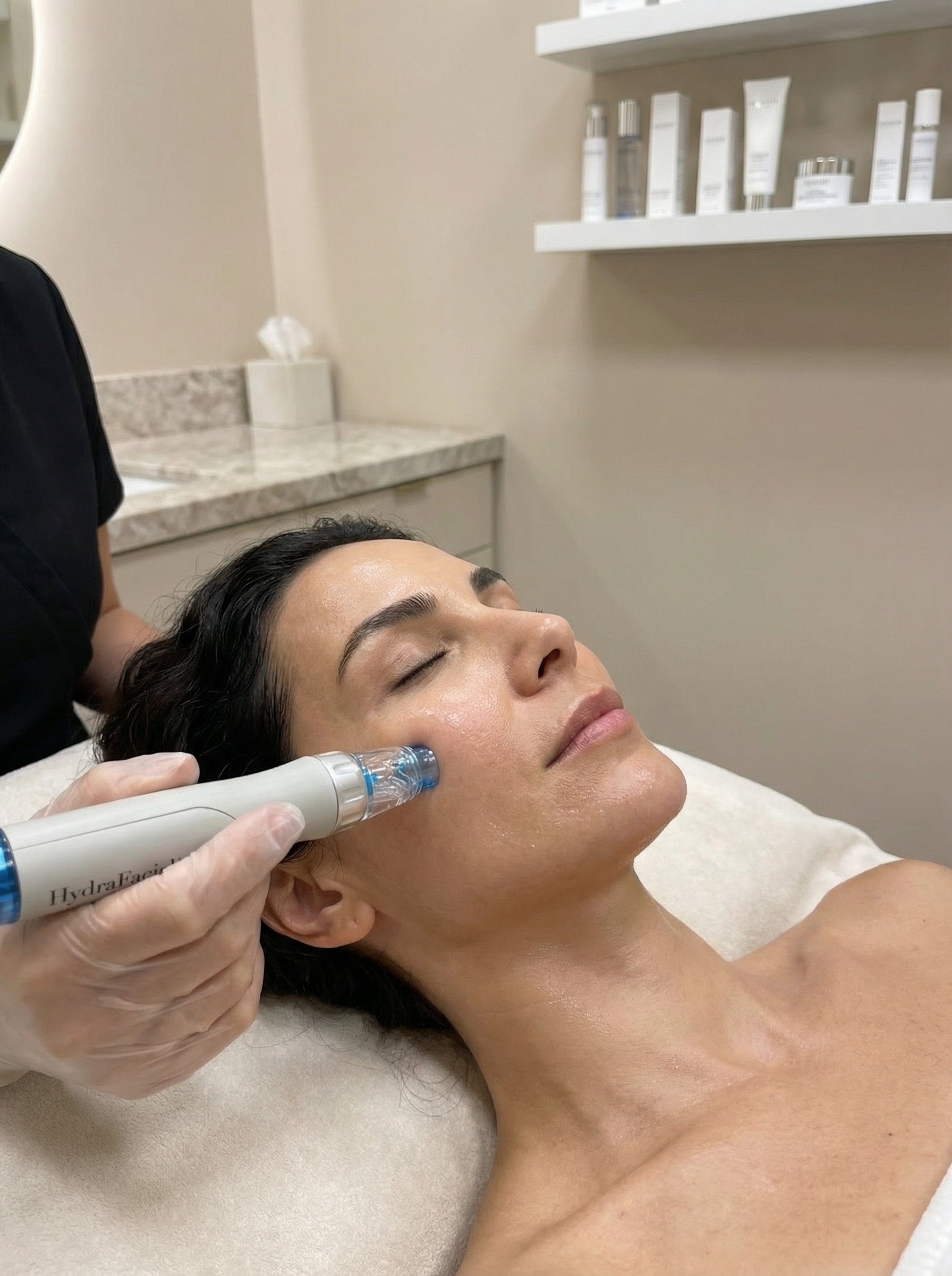 A woman lying with her eyes closed receives a cosmetic or dermatological treatment on her forehead using a handheld device by a professional wearing white gloves.