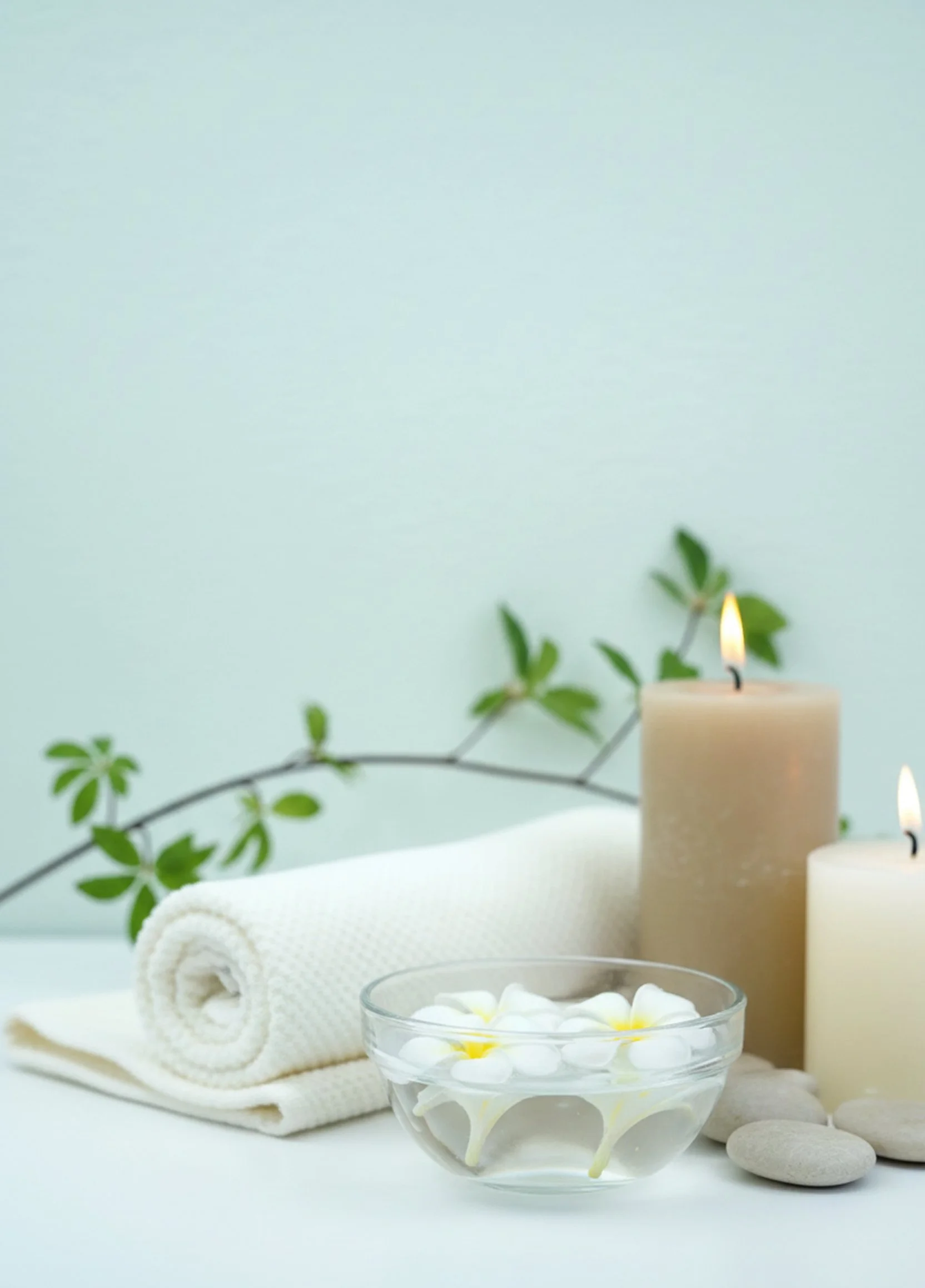 Spa setting featuring white rolled towel, glass bowl with white flowers, two lit candles, smooth stones, and green leafy branch.