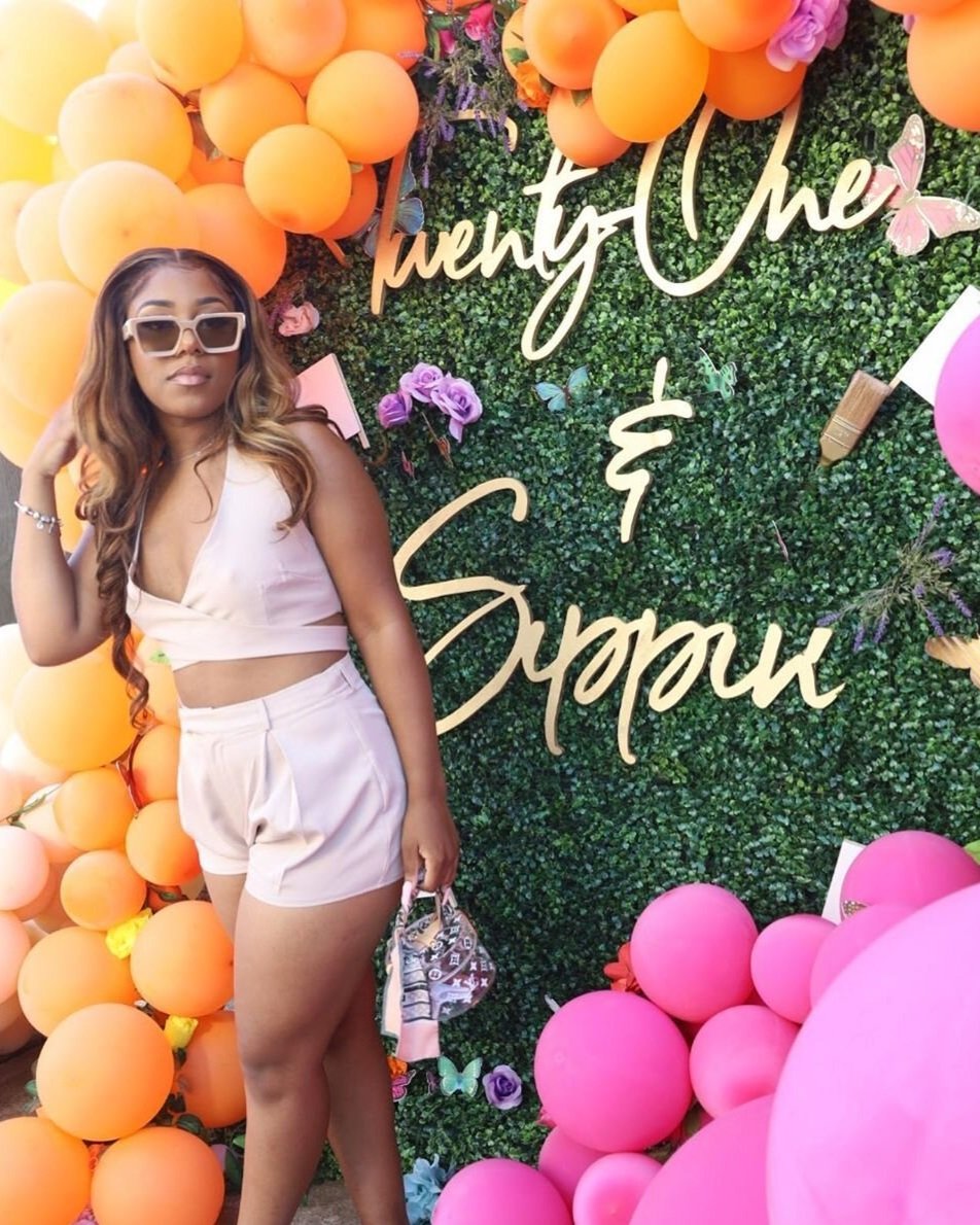 A woman in light pink clothing posing in front of a colorful backdrop of orange and pink balloons, green foliage, and decorative flowers with words and butterflies.