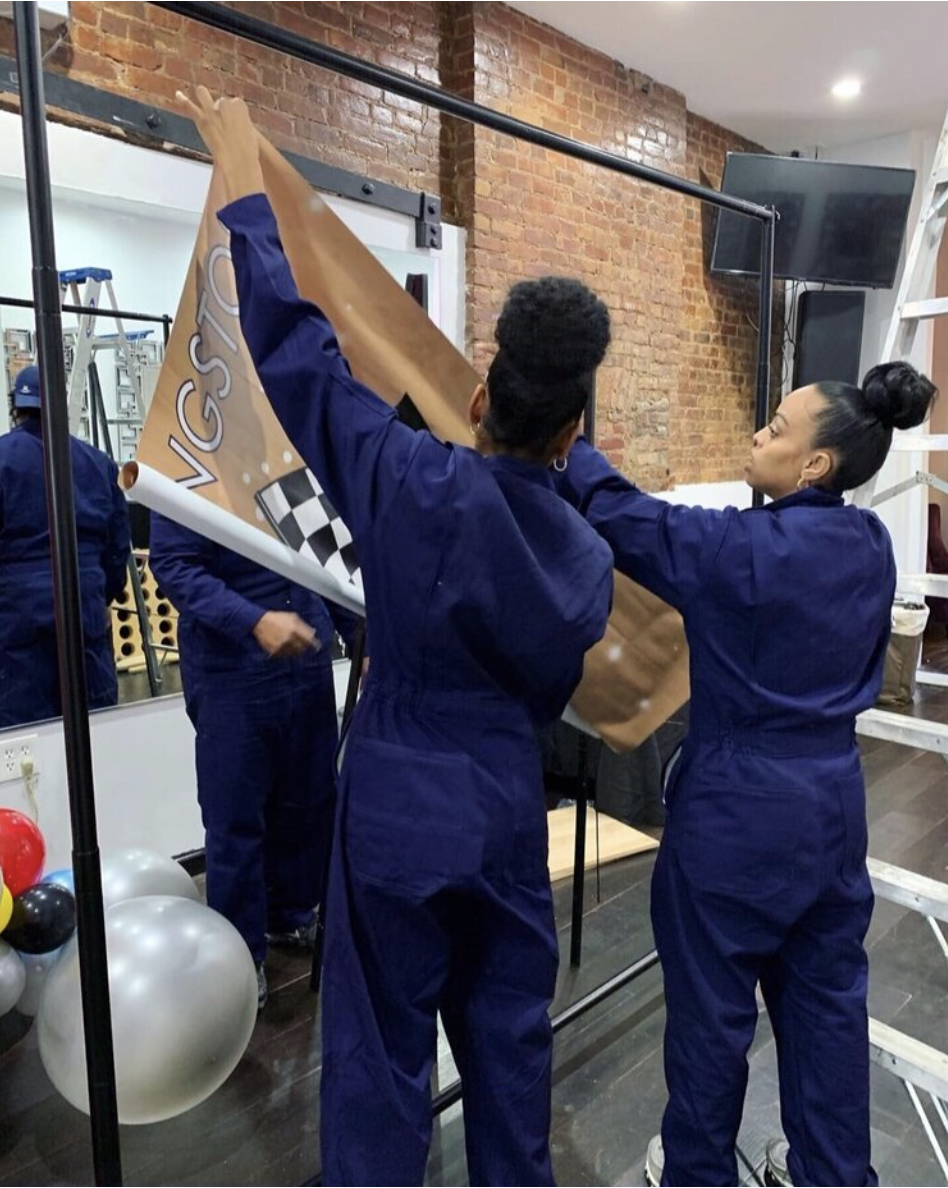 Two women in blue coveralls are hanging a large banner with checkered flags and racing symbols. They are in an indoor space with a brick wall, a mirror, and balloons nearby.
