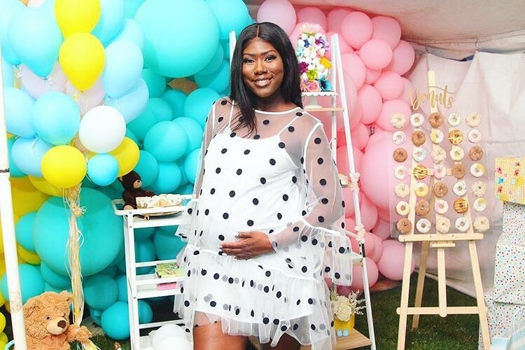 A smiling pregnant woman wearing a white dress with black polka dots and a sheer top, standing in front of a colorful party backdrop with balloons, decorations, and a dessert display.