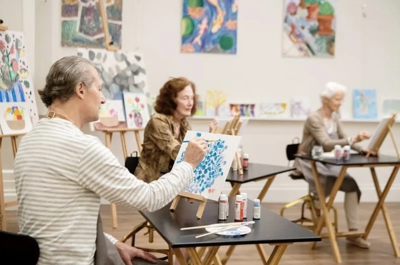 Older adults participating in a painting class in an art studio, each working on their own canvas with paints and brushes.