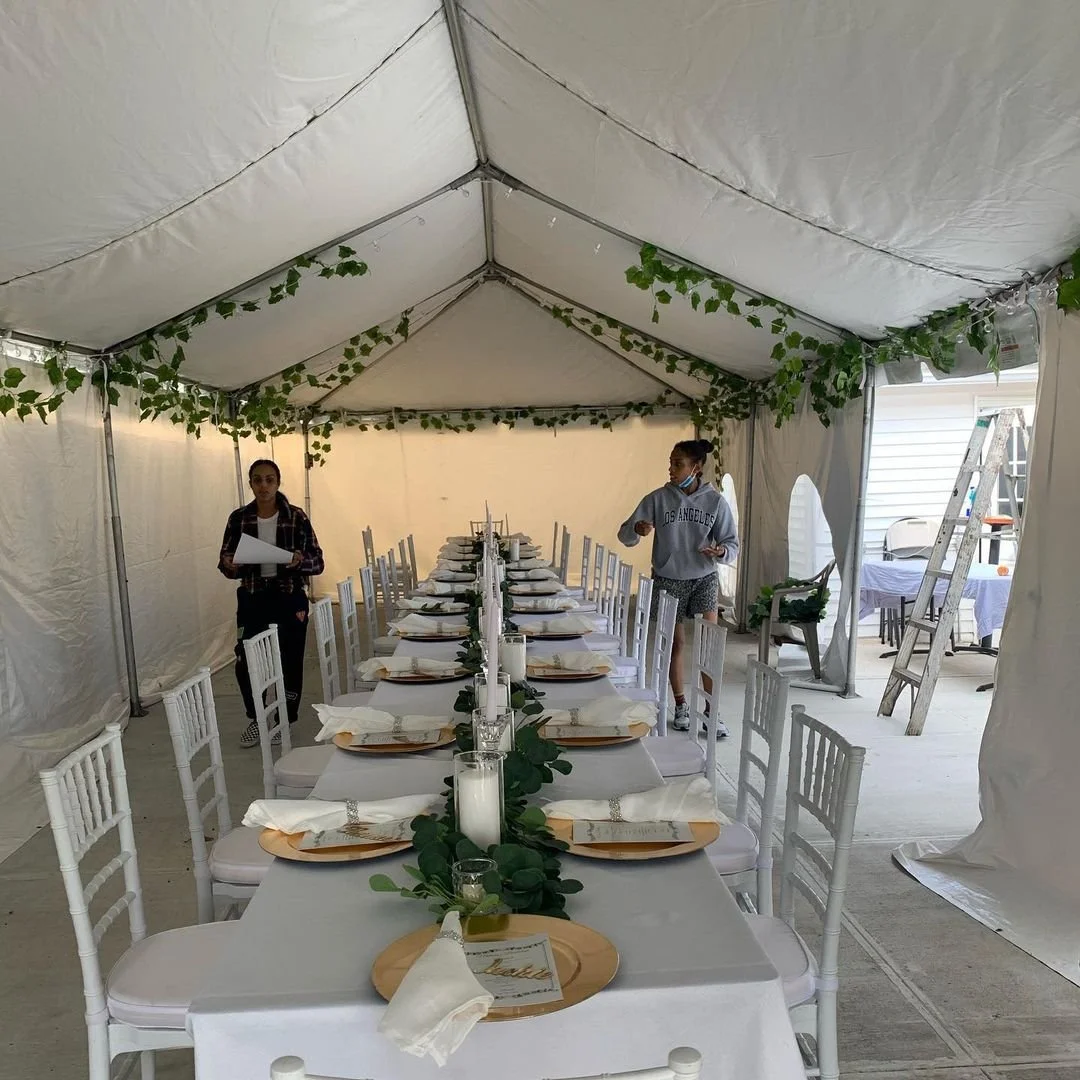 Long dining table set with white tablecloth, plates, napkins, and candles inside a decorated tent for an event or celebration.