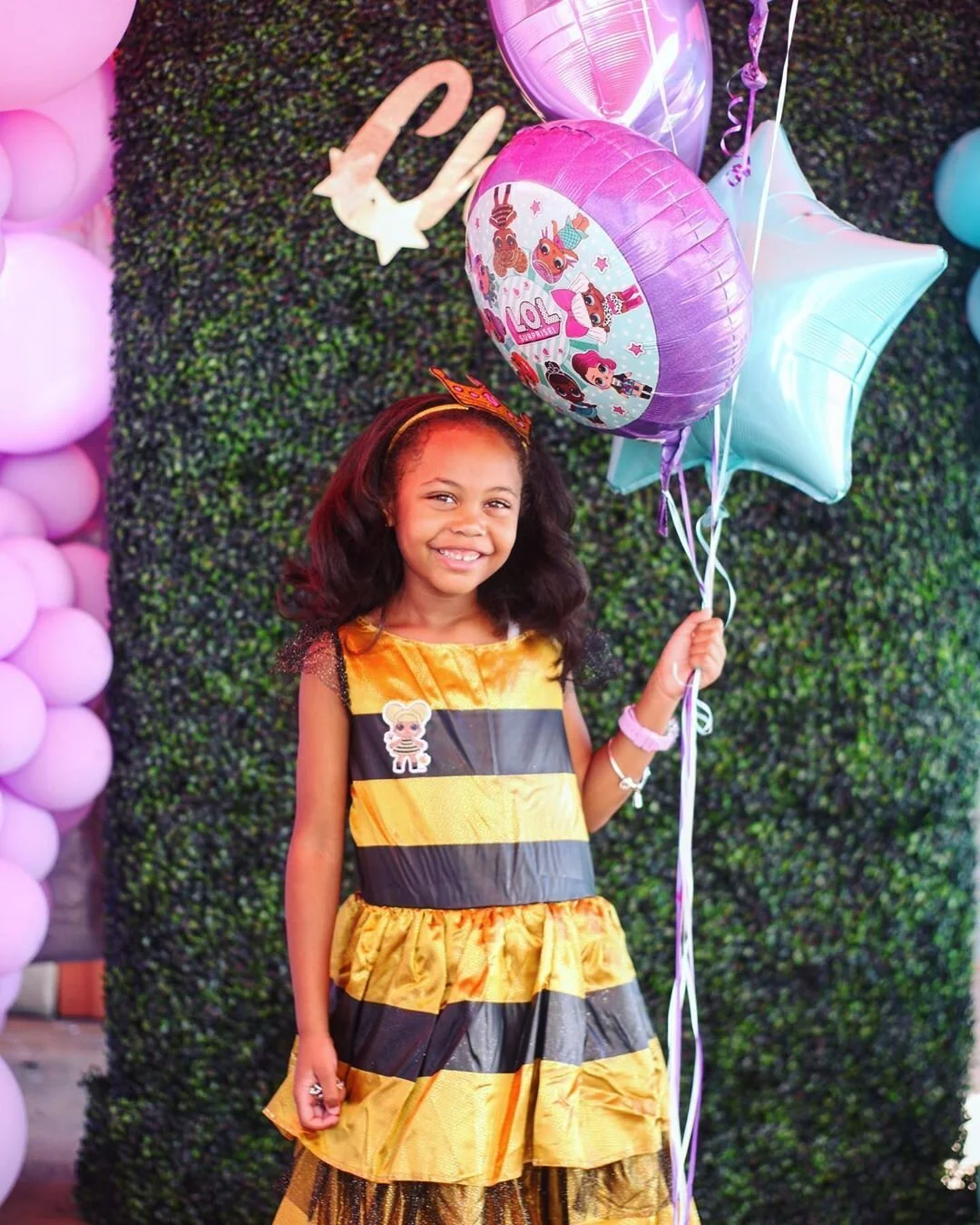 A young girl in a yellow and black striped dress holding balloons, standing in front of a green leafy background decorated with balloons and star-shaped cutouts.