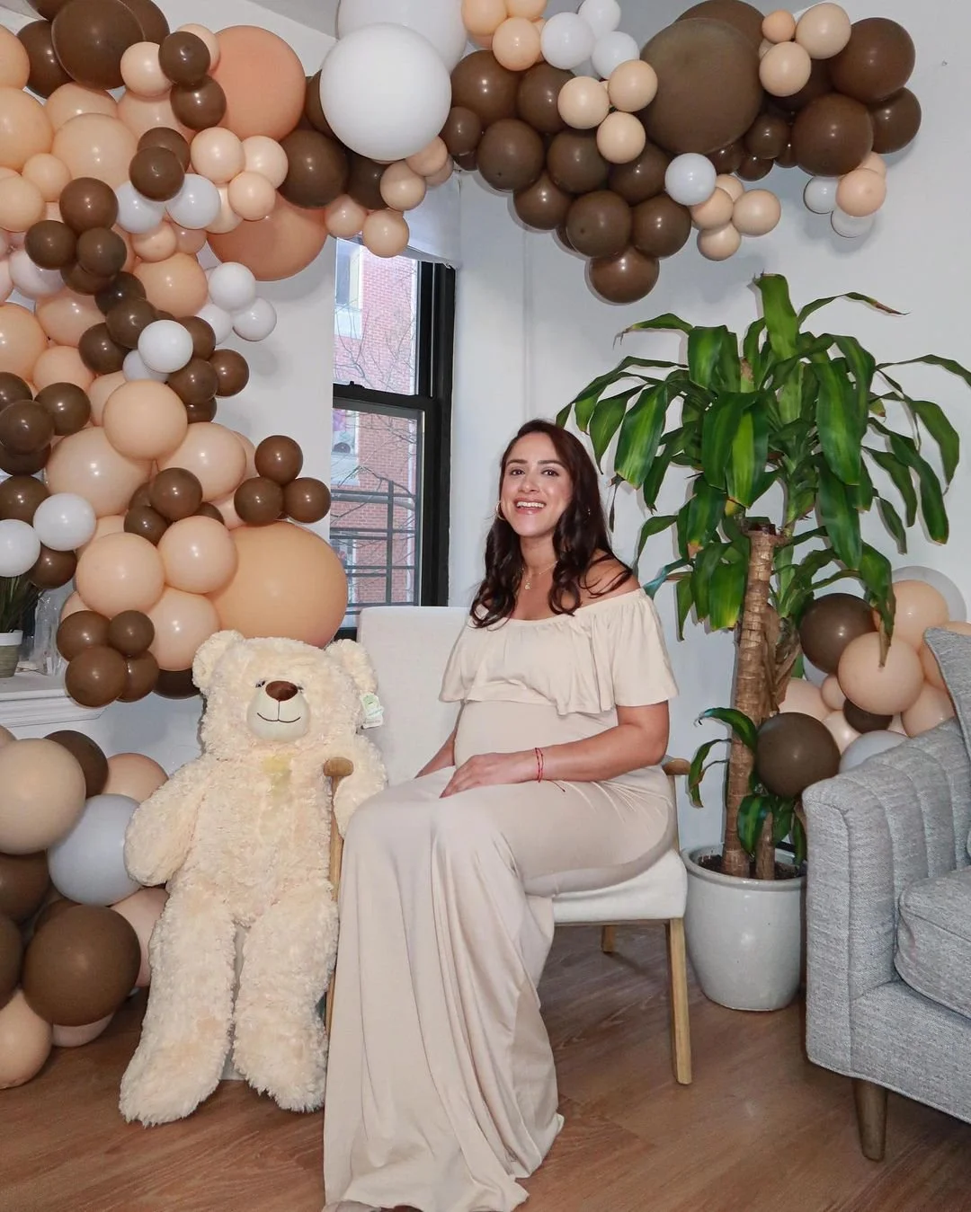A woman in a cream-colored dress sitting on a white chair with balloons and a teddy bear nearby. There is a large green plant, a window with a brick building outside, and various colored balloons on the wall and ceiling.