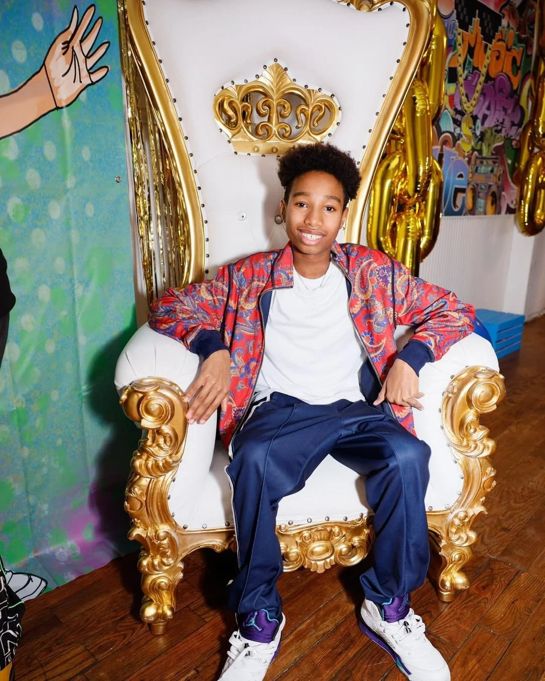 A young person with short curly hair sitting on an ornate white and gold throne with a high backrest, smiling at the camera. They are dressed in a white T-shirt, dark pants, a colorful patterned jacket, and white sneakers with purple accents. The background features colorful wall decorations and balloons.