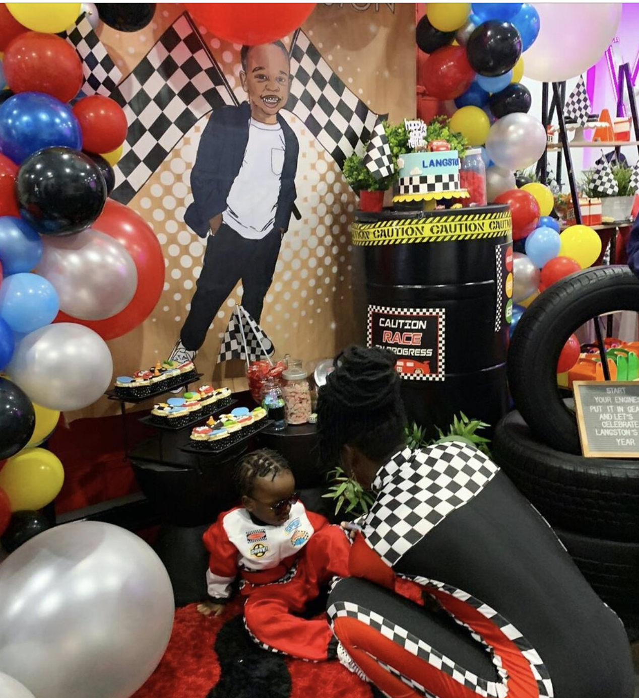 Children at a race car themed birthday party with balloons, checkered flags, race car decorations, and a cake with a race car design.