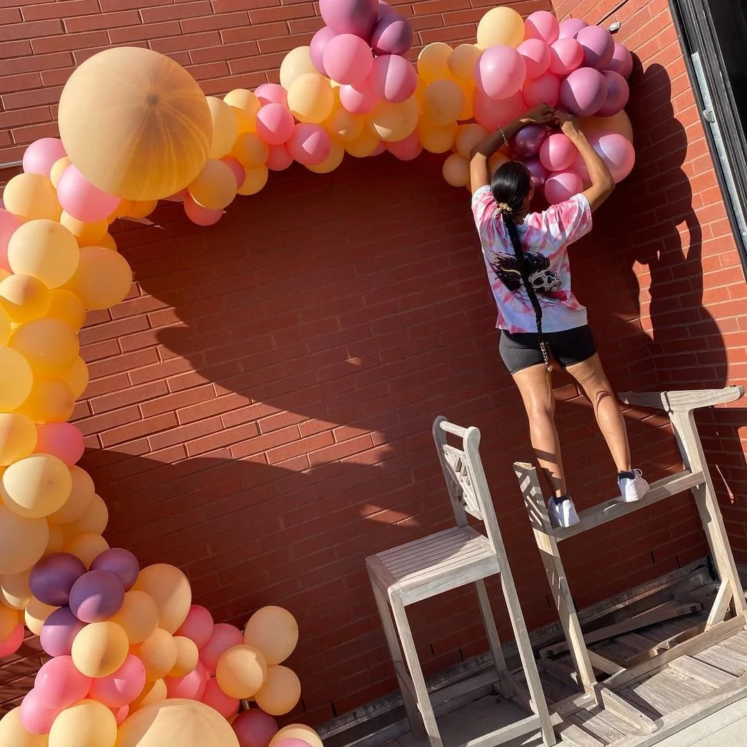 A woman arranging pink, purple, and yellow balloons on a brick wall with a large balloon arch. She stands on a wooden chair to reach the higher part of the balloon arrangement, wearing a colorful T-shirt and shorts.