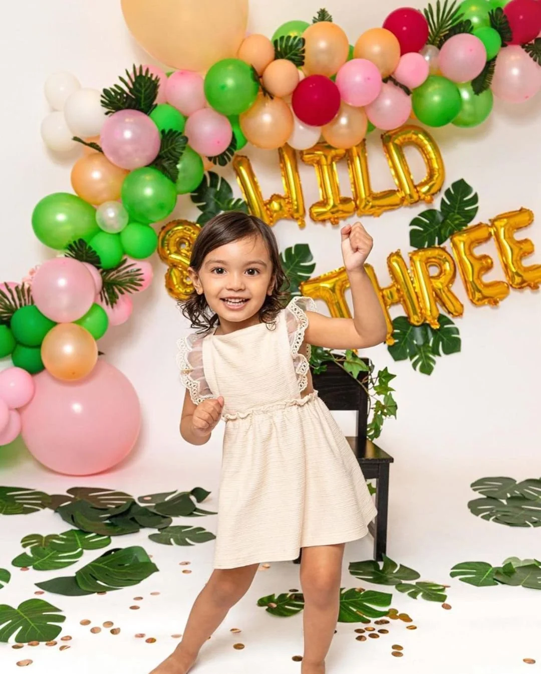 Young girl celebrating her birthday with a colorful balloon backdrop and gold letter balloons that spell out "Hello" and "Sisters." She is smiling, wearing a cream dress, and posing happily with one arm raised.
