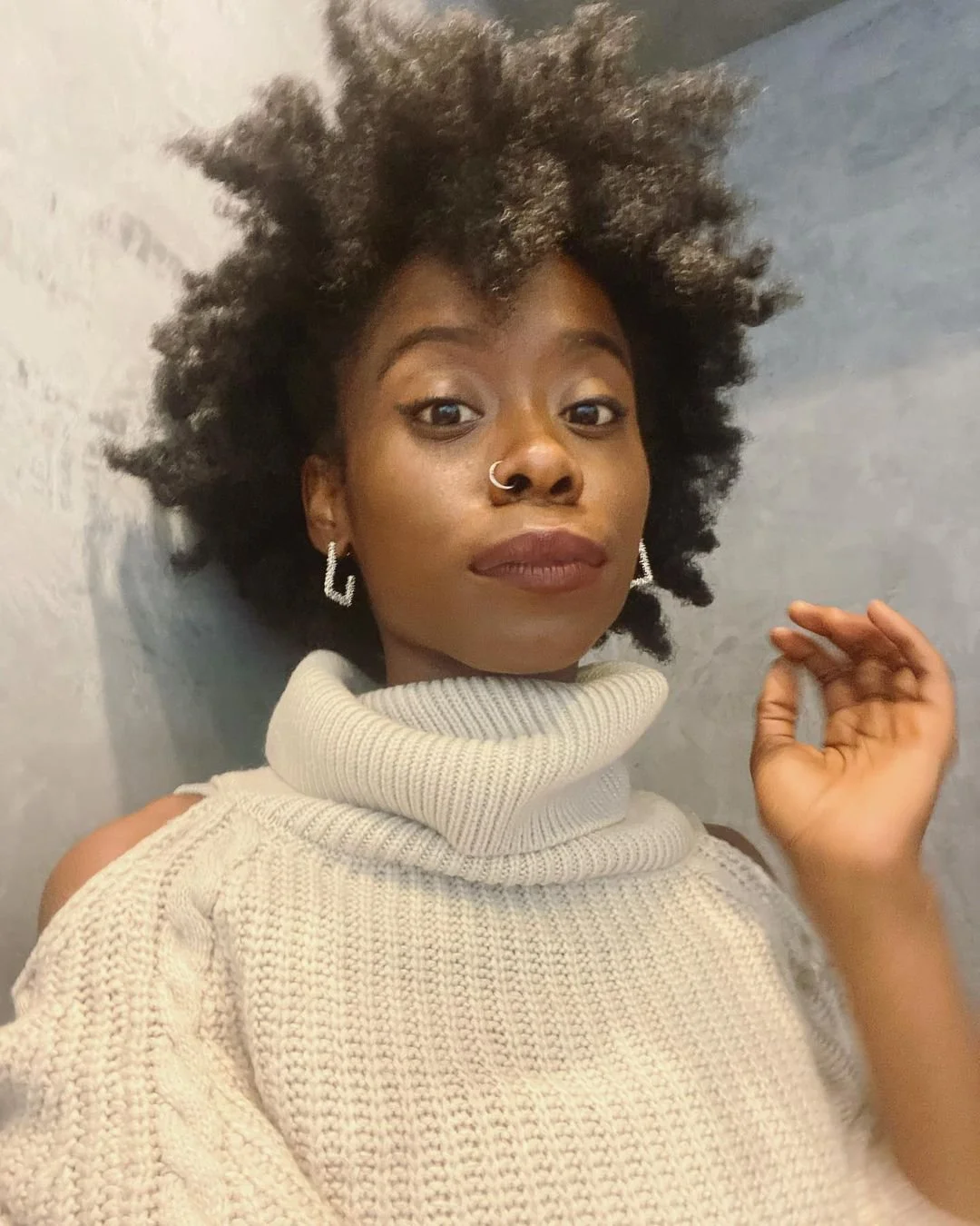 A woman with natural curly hair wearing a beige turtleneck sweater and silver earrings, looking at the camera against a gray background.