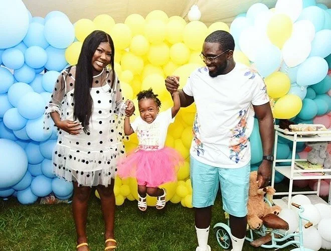 A family of three celebrating with a backdrop of yellow and blue balloons. The mother, father, and young daughter are holding hands and smiling, with the daughter jumping joyfully. The mother wears a polka dot dress, and the father wears a white T-shirt and turquoise shorts. The daughter is in a pink tutu dress with sneakers. There are toys and decorations on a table to the side.