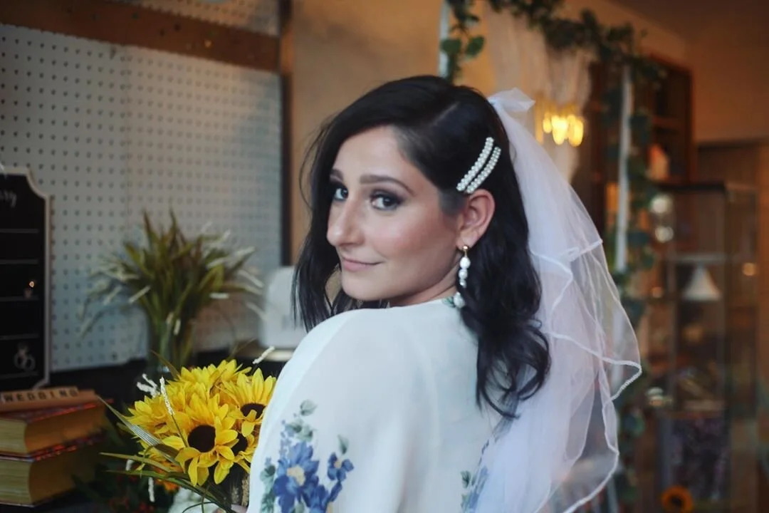 A woman with black hair, wearing a white veil and floral dress, holding a bouquet of sunflowers, in a warmly lit indoor setting.