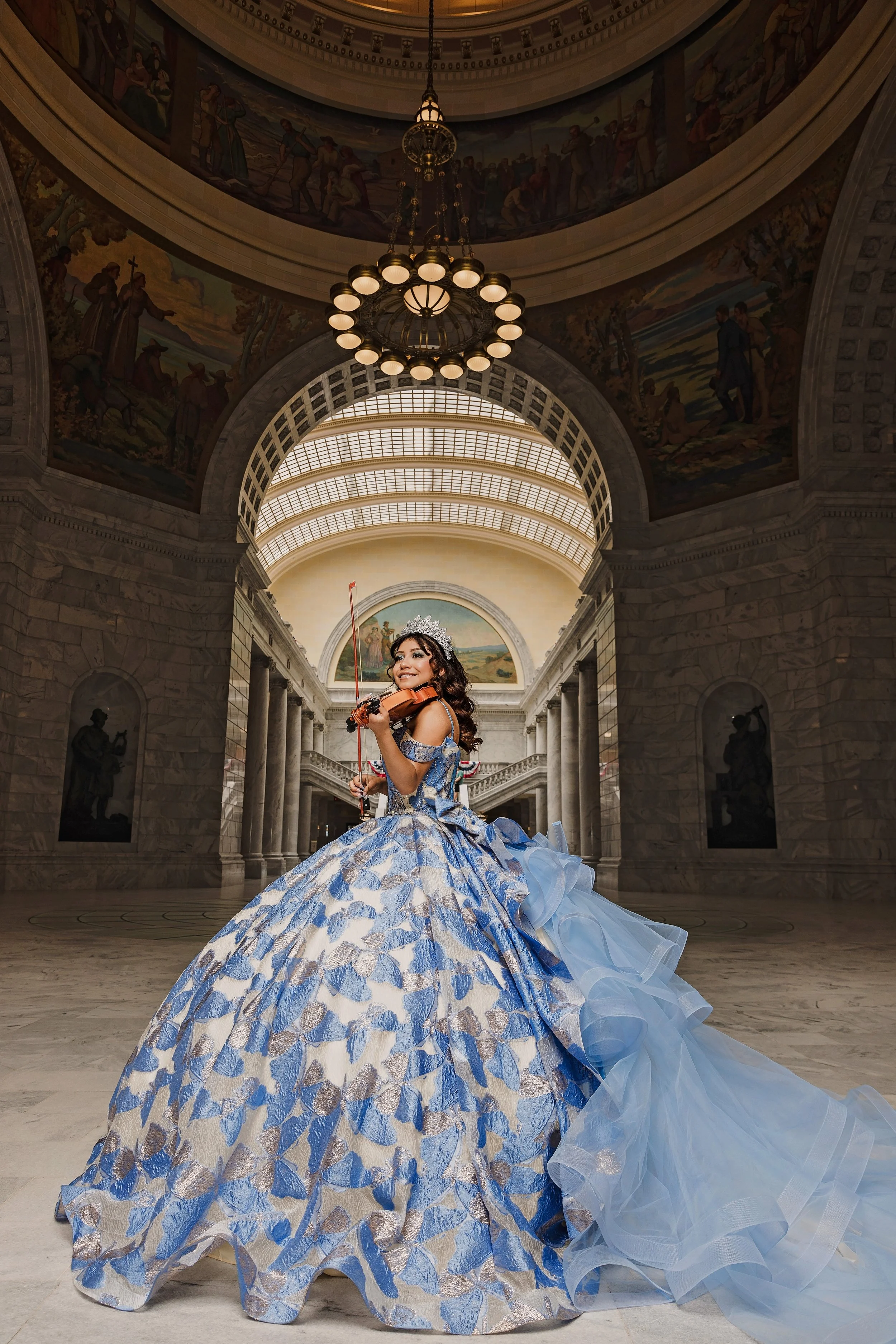 Capture the elegance of your Quinceañera with Once Upon a Quince, Utah’s premier Quinceañera photography and videography service. This stunning image showcases a young woman in a luxurious blue and silver gown, playing the violin under a grand chande