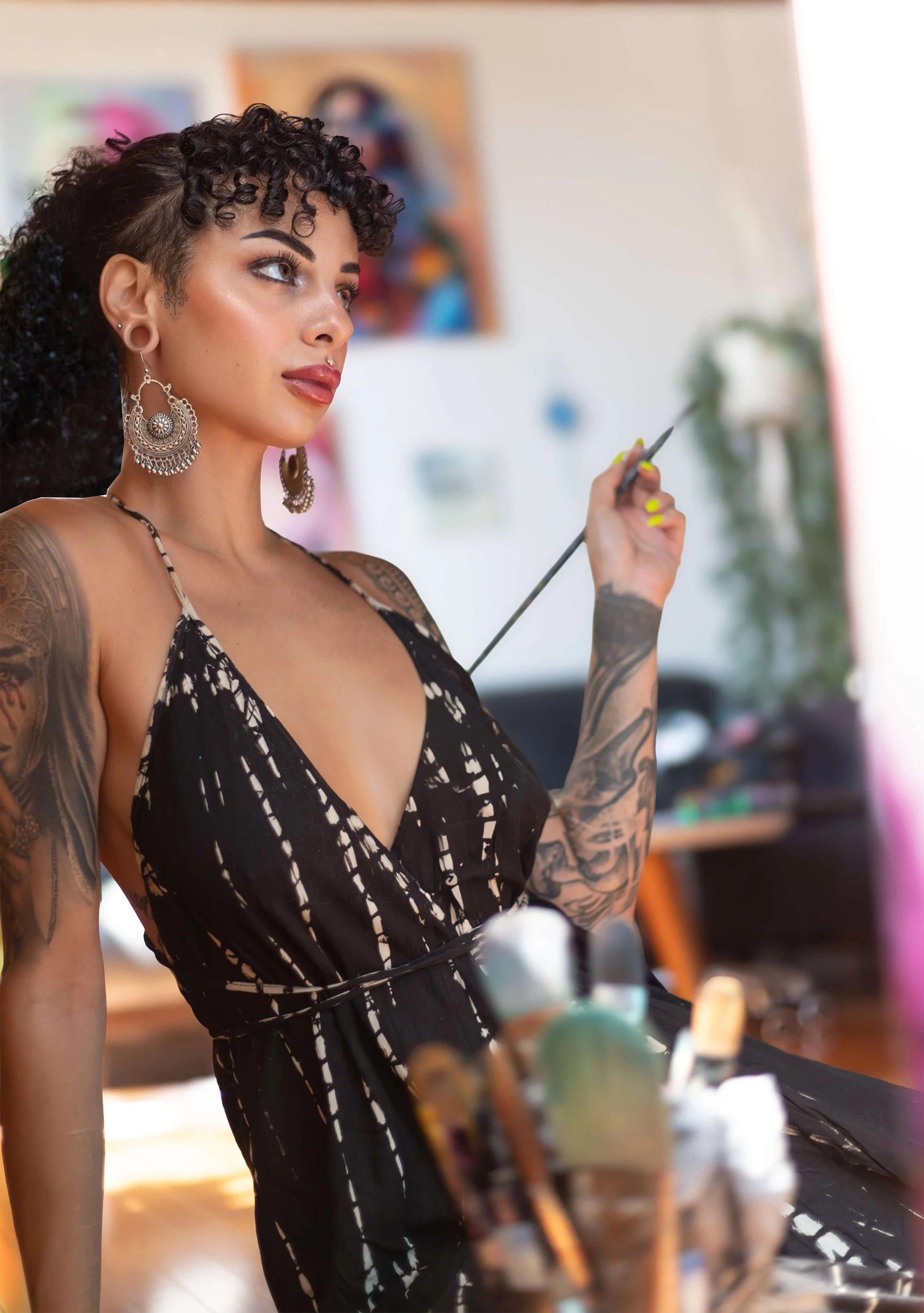 A woman with tattoos and large earrings, wearing a black and white patterned dress, holding a paintbrush or stylus, sitting near an artist's palette in a studio or creative space.