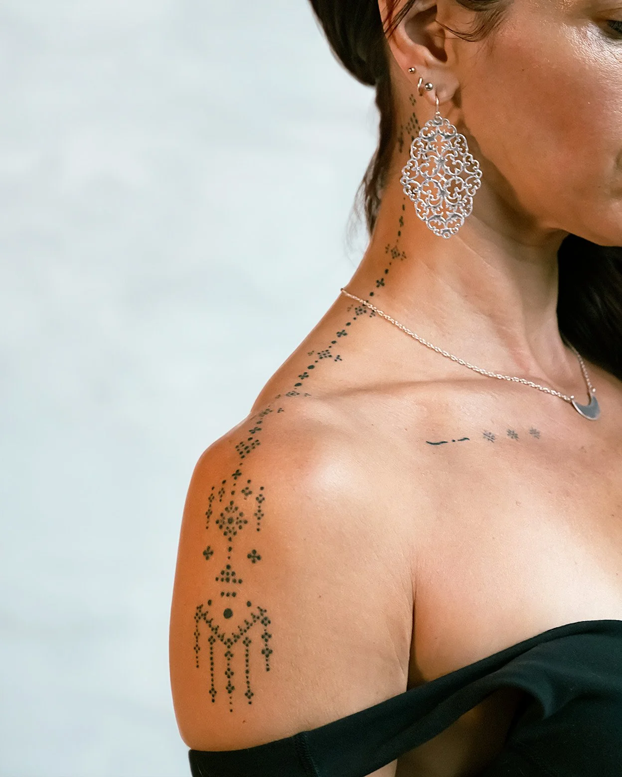 Close-up of a woman's shoulder and neck, showing multiple tattoos including a string of small symbols along her neck and shoulder, large ornate earrings, a necklace, and a portion of her black off-the-shoulder top.
