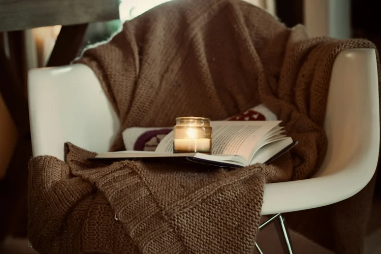 A cozy chair with a knit blanket, lit candle, and open book, inviting reflection and self-care
